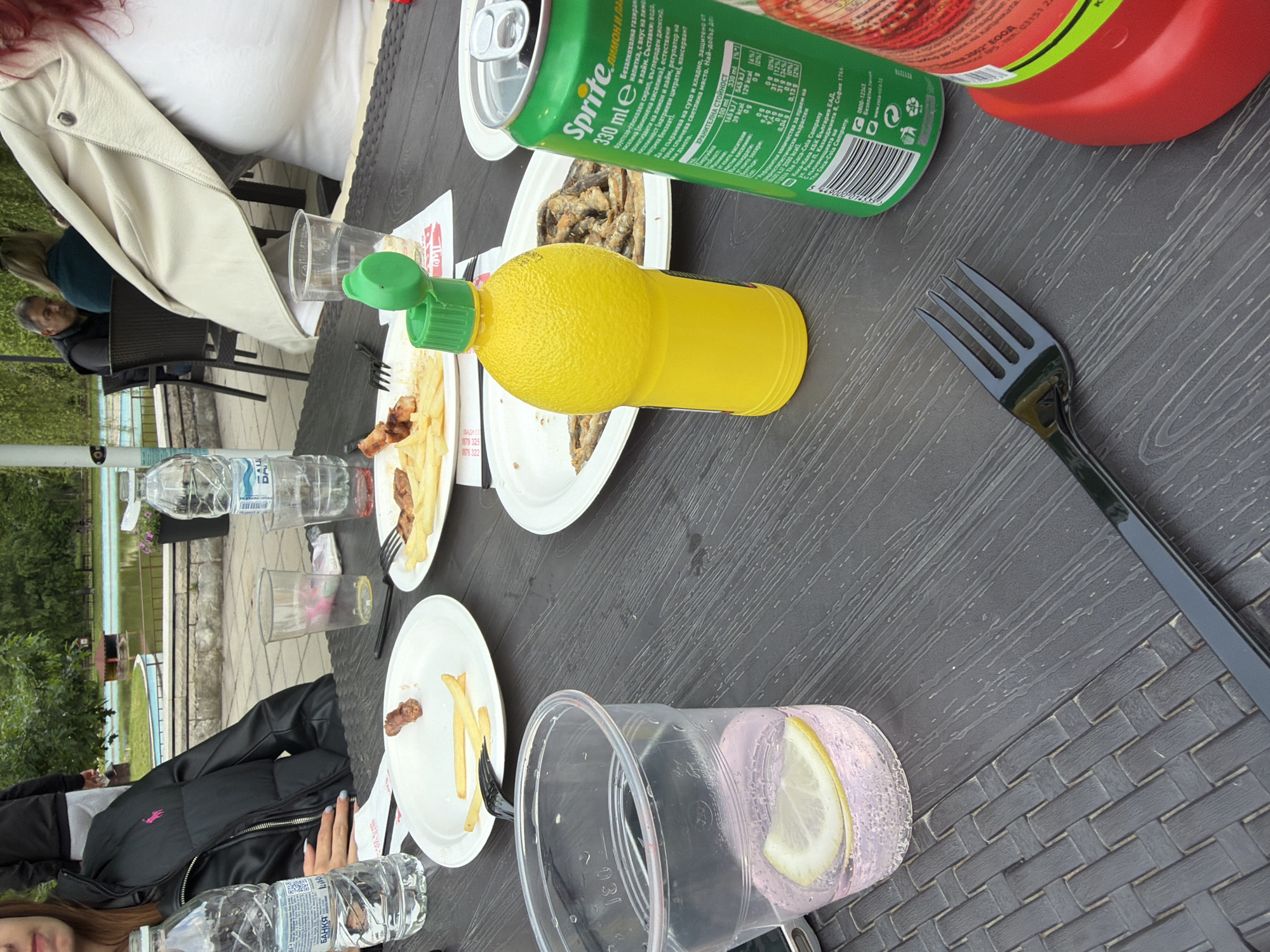 The image depicts an outdoor dining scene at what appears to be a casual restaurant or cafe, likely in Etropole, Bulgaria. A dark, textured table is laden with meal remnants and various beverages. Several white plates contain french fries and small pieces of fried fish or meat, along with black plastic forks.
On the table are multiple plastic bottles of "БАНКЯ" (Banka) brand water. A green Sprite can (330 ml) is prominent, with Bulgarian text indicating "ЛИМОН И ЛАЙМ" (Lemon and Lime) flavor, nutritional information, and "The Coca-Cola Company." A bright yellow lemon juice squeeze bottle and a red ketchup bottle are also present. One clear plastic cup holds a pink, bubbly drink with lemon slices.
In the foreground, a woman with red hair is partially visible, wearing a white top and a light-colored jacket. Another person, wearing a black jacket, is seated further down the table. In the background, a man with grey hair can be seen sitting at an adjacent table. The outdoor setting includes a blue body of water, possibly a swimming pool or pond, surrounded by greenery and stone landscaping. A piece of paper on the table displays Bulgarian text including "ОБАДИ СЕ" (Call), phone numbers "0878 322", "8879 325", and "2002 FOOD". The time appears to be daytime, suitable for outdoor dining.