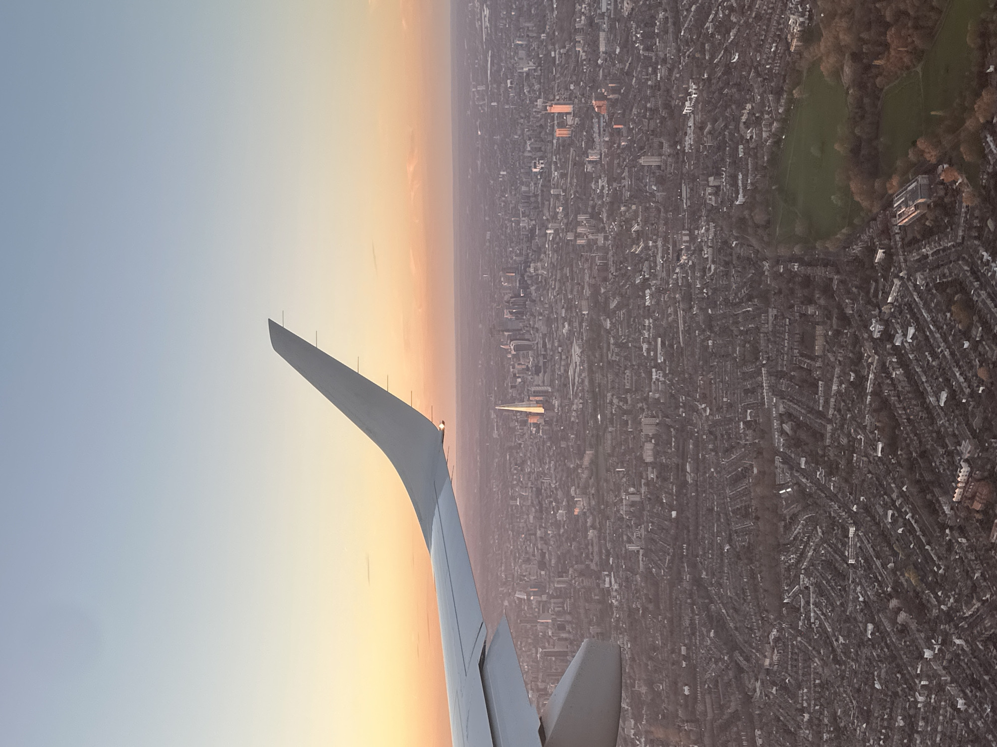 An aerial view from an airplane window shows an aircraft wing with a cityscape in the background. The sky transitions from blue to orange hues near the horizon. The cityscape is dense with buildings and street patterns. In the distance, a green park area is visible. Several tall buildings are scattered within the urban landscape, including a prominent pyramid-shaped skyscraper. The location appears to be the city of London, United Kingdom.