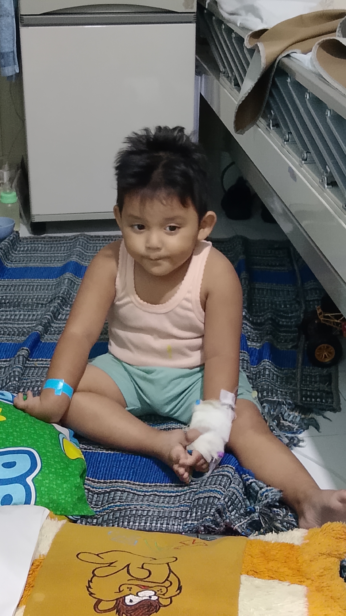 A young child with dark hair and olive skin sits cross-legged on a patterned blanket on the floor. The child is wearing a light pink tank top and light blue shorts. The child's right arm is bandaged, with tubing visible. A blue bandage is on the child's left arm. A green pillow with blue lettering is to the child's left, and a yellow pillow with a cartoon character is in the foreground. In the background, a white refrigerator and a metal bed frame are visible. A black toy car is on the blanket to the right. The scene appears to be indoors.