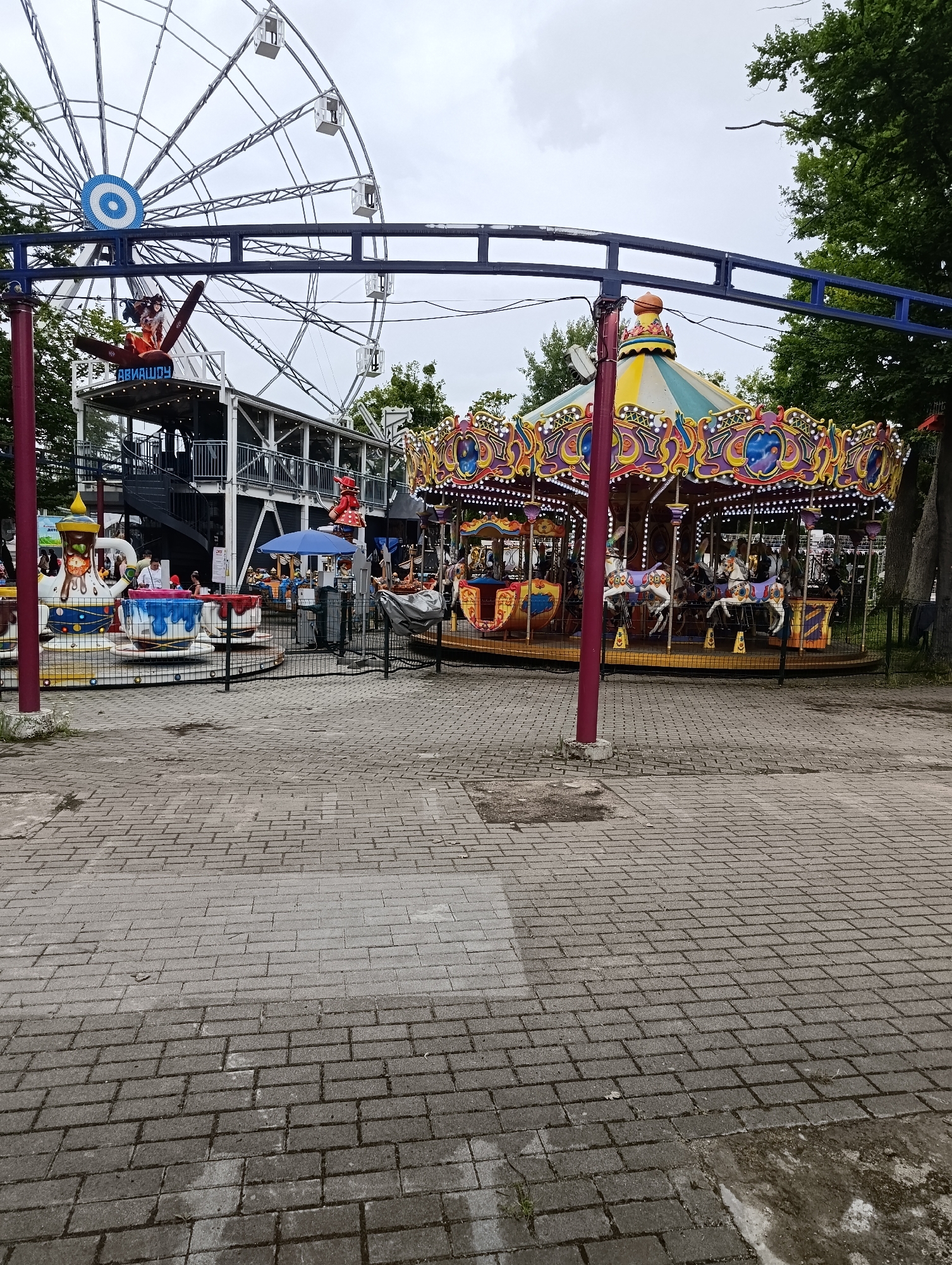 This is a daytime, medium-shot, eye-level, exterior, wide-angle photograph of an amusement park in Kaliningrad, Russia. In the foreground, a paved walkway made of light-gray rectangular bricks leads into the park. To the left of the walkway is a small, white-and-blue teacup ride with several teacups. To the right of the teacup ride is a carousel featuring colorful horses and ornate decorations. In the background, a large Ferris wheel with white cabins is visible, along with trees and a cloudy sky. Several people are present in the park, some standing, some appearing to be riding the attractions. The overall atmosphere suggests a recreational, family-friendly environment.