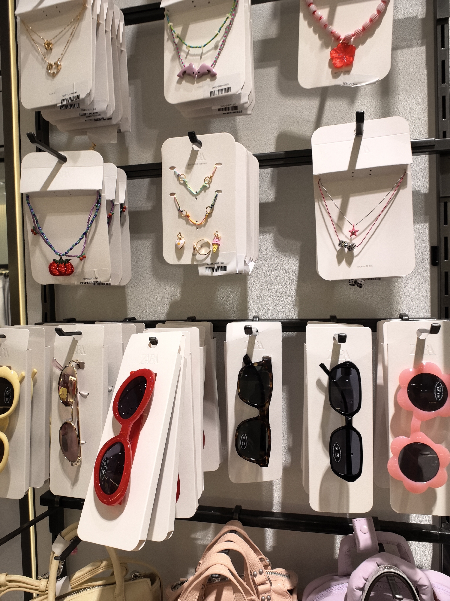 The image shows a retail display of various fashion accessories, including necklaces and sunglasses, against a textured gray wall. The items are neatly arranged on white cardboard backing and hung on black horizontal racks. In the top section, several necklaces with colorful beads and pendants like hearts, flowers, and dolphins are displayed. Below these, a selection of sunglasses is presented. These include pairs with tortoiseshell frames, black frames, and a novelty pair with pink flower-shaped lenses. The lighting is even, suggesting an indoor retail environment. The bottom of the image shows portions of handbags, with a light beige bag on the left and a pink bag on the right.
