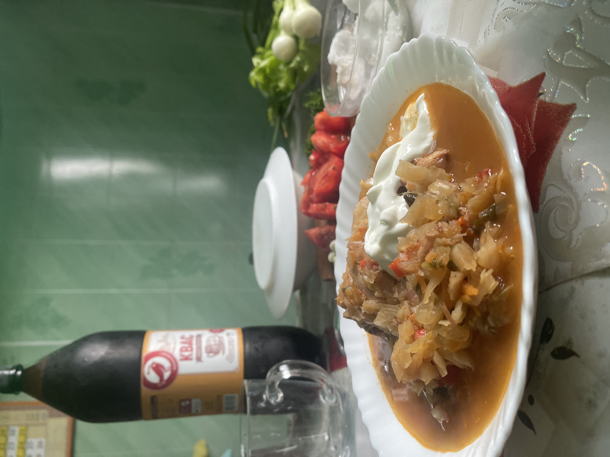 A plate of stew, topped with sour cream, sits on a table covered by a patterned tablecloth. In the background, a bottle of kvass, two glasses, chopped tomatoes, green onions, and a bowl of salt are visible. The scene is set in a kitchen with green walls. The city name "Красное" appears on the bottle of kvass.