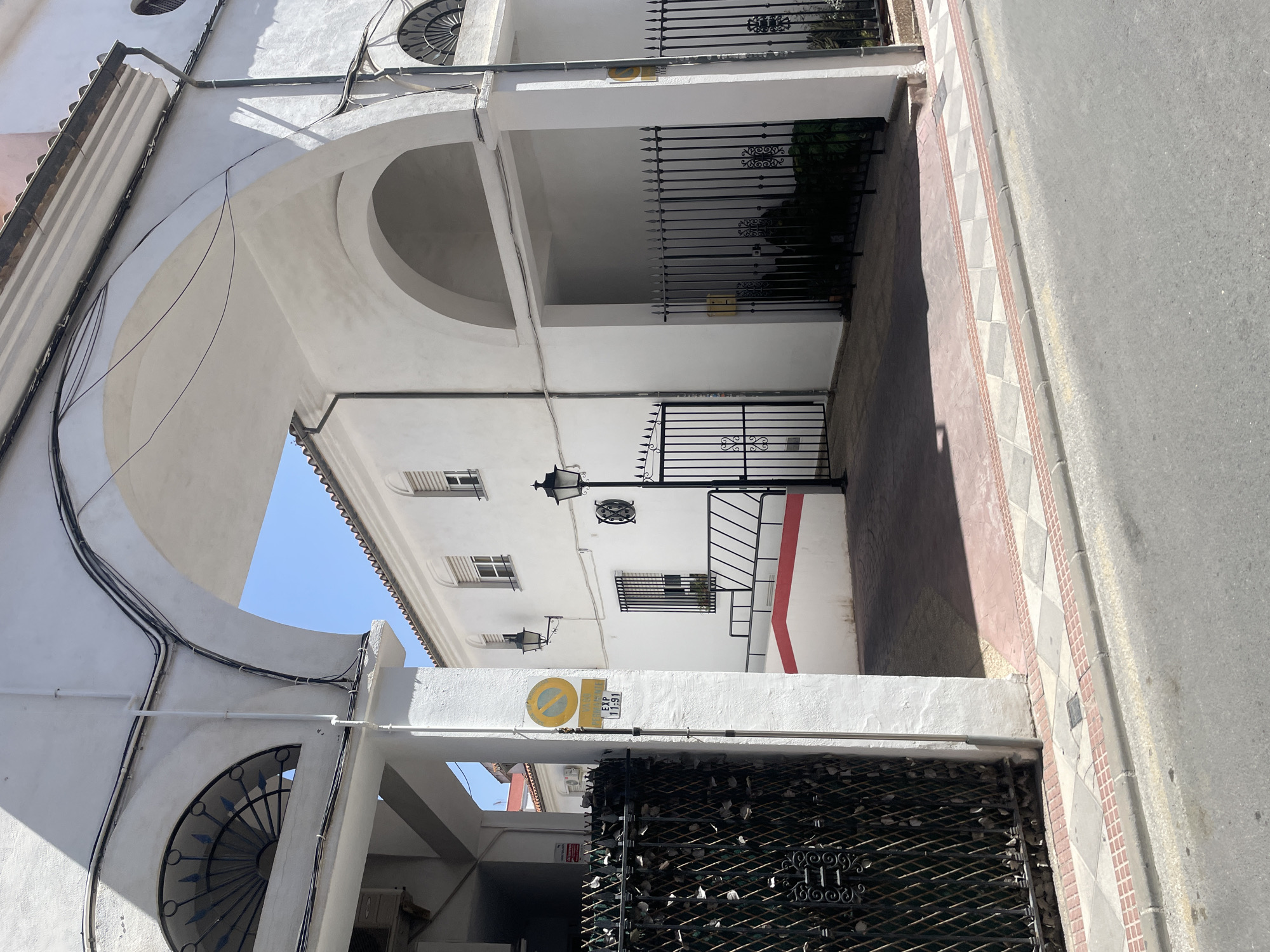 The image depicts a street scene in the city of Atarfe, Spain, focusing on the facade of a white building with arched structures. The building features white walls, white arched passages, and a tiled roof. In the foreground, a paved street is visible with a sidewalk made of decorative tiles.
On the left side of the image, a black wrought-iron gate is prominent, partially obscured by a white column. A circular yellow sign with a red border and a diagonal red line through it, indicating a prohibition (likely parking), is attached to this column. Below it, a rectangular white sign with black text "EXP 11-91" is also affixed. Adjacent to the gate, an air conditioning unit is visible on the building's exterior.
The central part of the image shows an arched walkway leading into a courtyard or interior space. Within this space, a black wrought-iron gate is closed. To the right of this gate, another black wrought-iron fence with decorative finials encloses a small area with greenery. A street lamp with a black metal post and a lantern-style fixture stands to the left of the central archway.
The building to the right of the archway has several windows with white shutters. Another street lamp with a similar design is mounted on this wall. The architecture suggests a Mediterranean style. The sky is clear and blue, indicating a sunny day. The lighting casts distinct shadows on the ground and walls. The overall impression is one of a quiet urban street with traditional architectural elements.