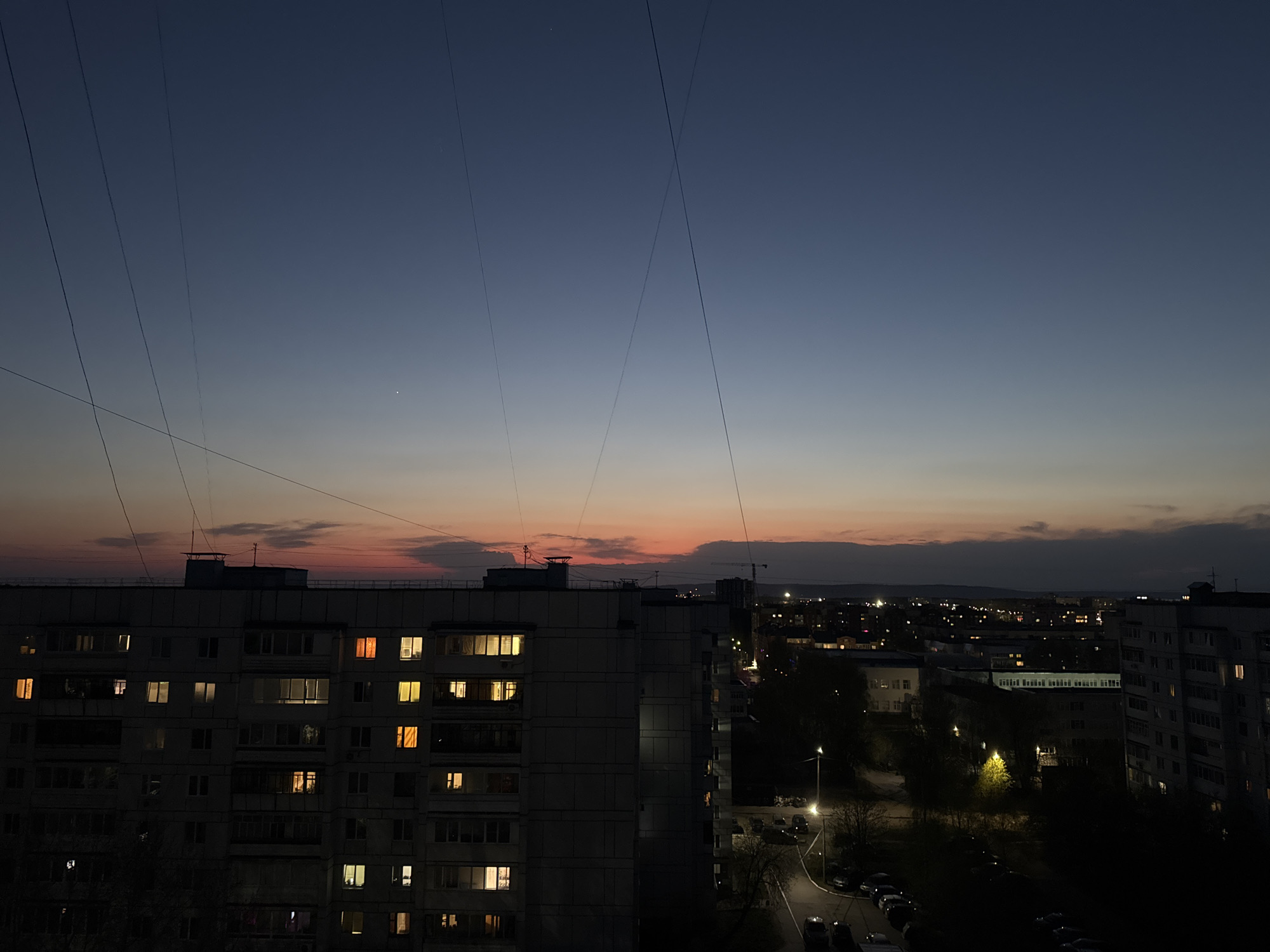 The image depicts a cityscape at dusk in what is likely Chaikovsky, Russia. The sky transitions from deep blue at the top to shades of orange and pink near the horizon. Several thin wires stretch across the sky. In the foreground are apartment buildings, with a few windows illuminated. Further in the distance are city lights, trees, and more buildings. Cars are parked along the street. There are no people visible.