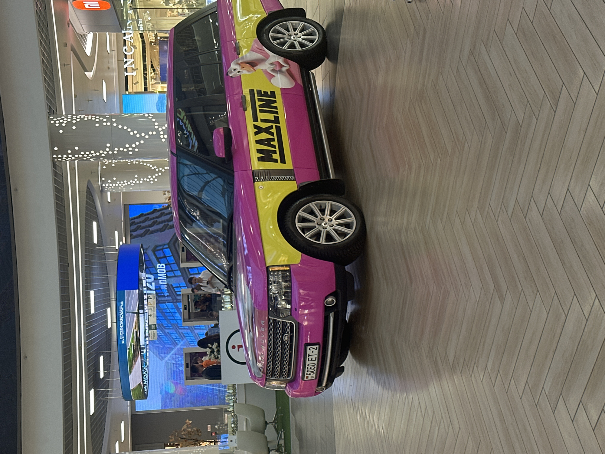 A pink and yellow Range Rover is parked indoors on a herringbone patterned wooden floor. The vehicle has the text "MAXLINE" in large black letters with a yellow outline on its side, along with a cartoon duck character wearing a pink shirt and making a heart shape with its hands. The license plate reads "5050 ET-2" and has a Belarusian flag sticker on the left. In the background, there are escalators, shops with signage like "INCANTI", and large digital displays showing urban scenes and text, some of which appears to be in Cyrillic. People are visible in the background near the escalators and shops. The environment suggests a modern shopping mall or exhibition space in Minsk, Belarus.