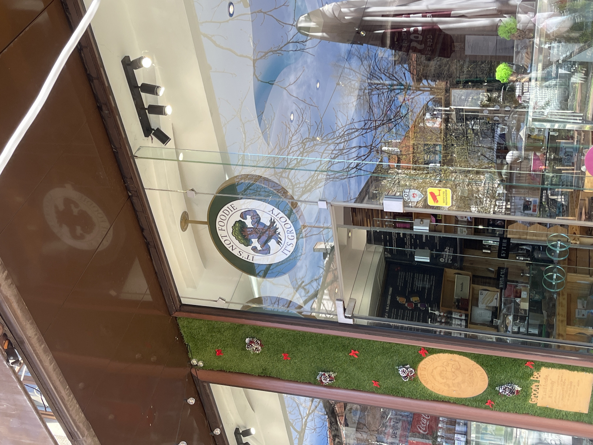 The image shows the storefront of "It's Not Foodie It's Grooty", a business in Sofia, Bulgaria. The storefront has a logo on the glass door. The window display includes artificial grass, decorative items, and signage. Reflections of the street and sky are visible in the glass.