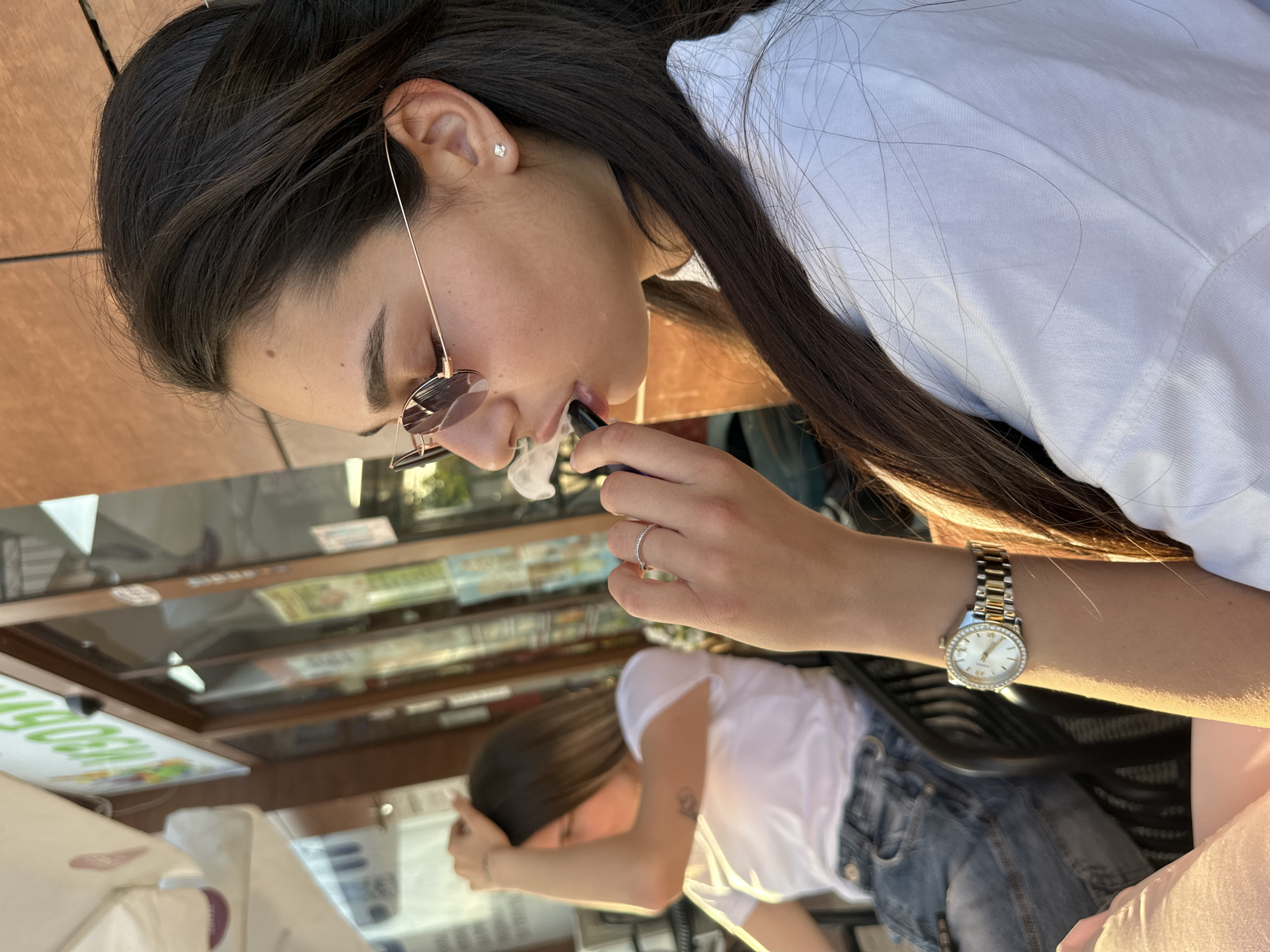 The image shows a woman with long, dark hair wearing sunglasses and a white shirt. She is holding a vaping device to her lips and exhaling vapor. She has a ring on her finger and a watch on her wrist. In the background, there is another person with dark hair wearing a white shirt and jeans, their face obscured. Behind them are shelves with various items. The scene appears to be outdoors, possibly at a market or cafe in Plovdiv, Bulgaria, suggested by the architecture and merchandise in the background.