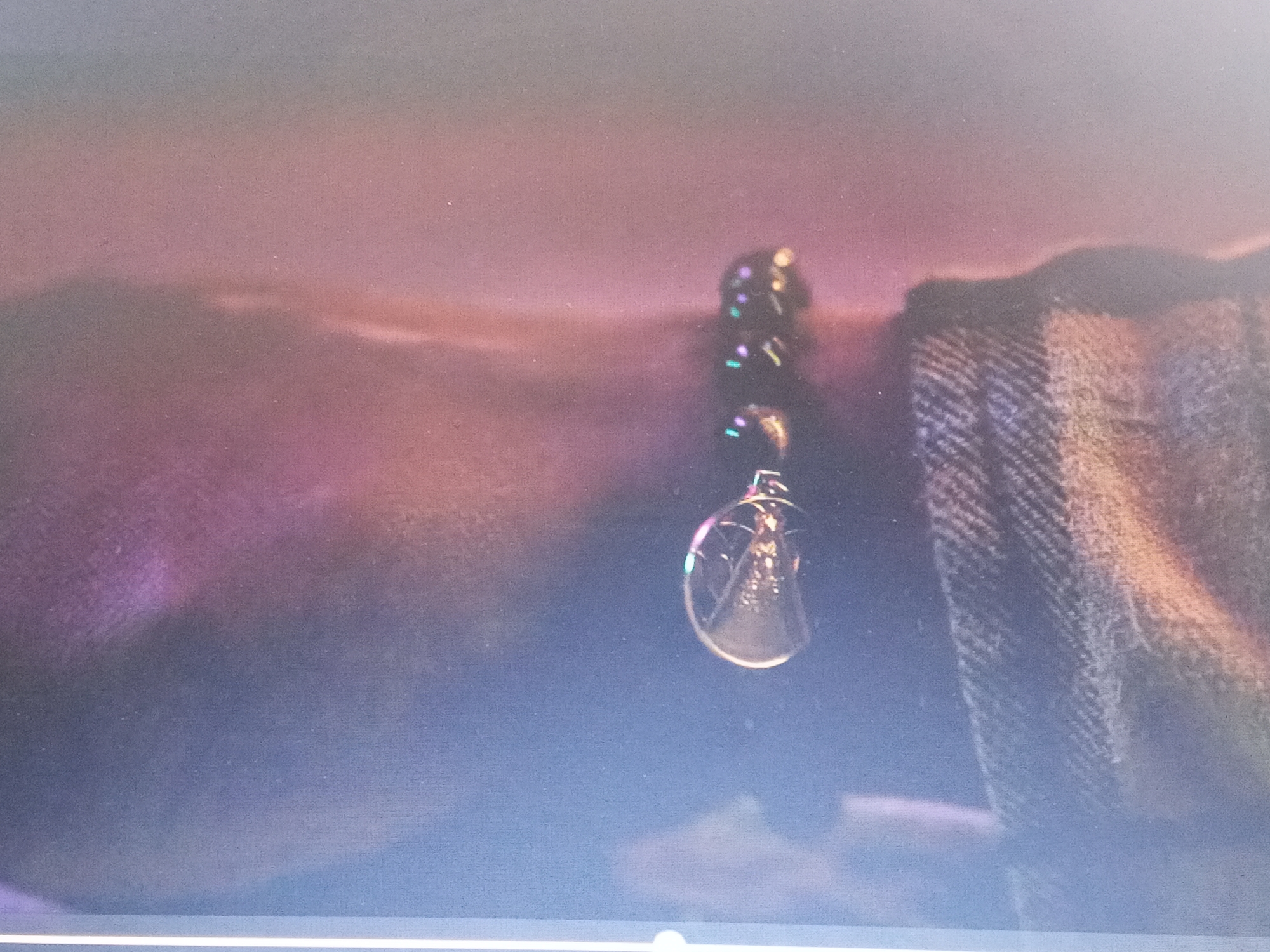 The image shows a close-up of a beaded bracelet with a pendant hanging from it. The bracelet is draped over what appears to be a denim or woven fabric, possibly clothing. The background features blurred purplish and pinkish tones. The pendant is circular and appears to contain a figure inside, possibly a small statue or design. The scene doesn't provide enough information to determine a location in Gwong, Nigeria or provide further details.