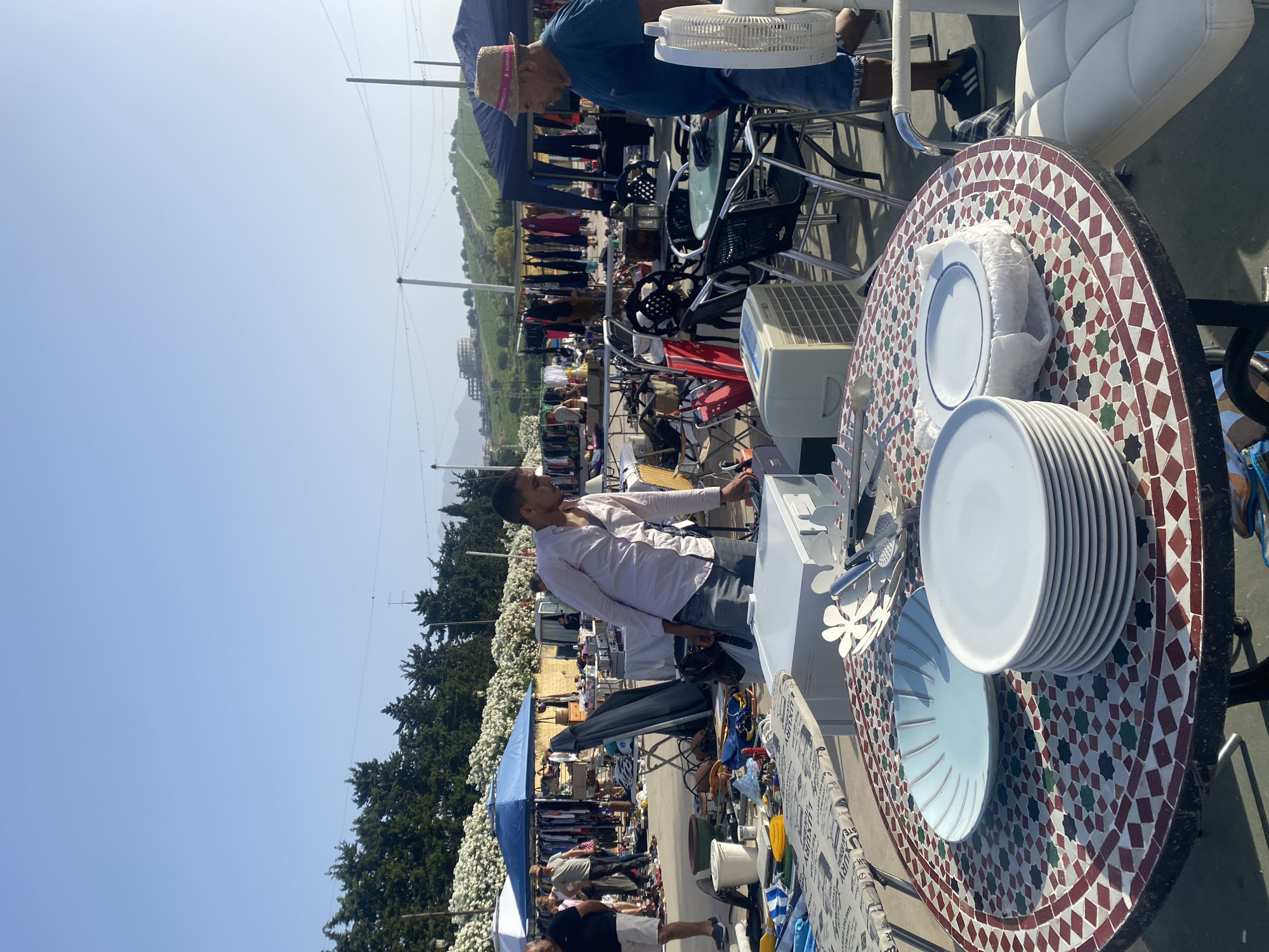This image depicts a flea market scene in Manilva, Spain, under a clear, bright sky. In the foreground, a round table with a mosaic tile top displays a stack of white plates, a light blue bowl, and a metallic serving utensil. To the right of the table, a white upholstered chair with a chrome frame is partially visible.
In the mid-ground, two men are interacting. One, wearing a light pink collared shirt and blue jeans, appears to be examining items on a table or stall. The second man, wearing a blue t-shirt, denim shorts, and a straw hat, is standing near a table with chairs, looking towards the items on display. A white portable fan is positioned near him.
The background reveals rows of stalls with clothing and various goods displayed under blue and dark blue awnings. Several people are browsing these stalls. Lush green trees with white flowers line the perimeter of the market area, and in the distance, a hillside with buildings and vegetation is visible. Power lines and utility poles are also present. The overall setting suggests an outdoor market on a sunny day.