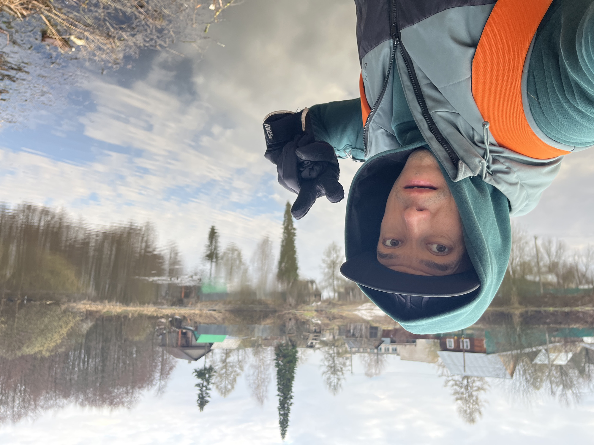 A man in a teal/green hooded jacket and a grey/black padded vest with orange straps is captured in what appears to be a selfie or direct portrait. He is wearing a dark cap under his hood and black gloves, with the text "2AV" visible on the left glove's wrist strap. The man is looking directly at the camera with an open mouth, displaying a surprised or playful expression, and making a hand gesture, possibly a "shaka" sign, with his left gloved hand.
The image is intentionally inverted, creating an optical illusion where the man and the distant landscape appear to be reflected in a body of water that acts as the primary foreground. The upper portion of the image, representing the water, reflects a cloudy sky and the inverted distant shore. The lower portion, representing the sky, reflects the upright distant shore.
The scene depicts a rural or semi-rural environment, likely during late winter or early spring in Chaikovsky, Russia, given the bare trees on the distant banks. Scattered wooden houses, some with green roofs, are visible along the shoreline. The sky is bright with light clouds, suggesting daytime. The overall impression is playful, utilizing the reflective properties of the water to create an unusual perspective.