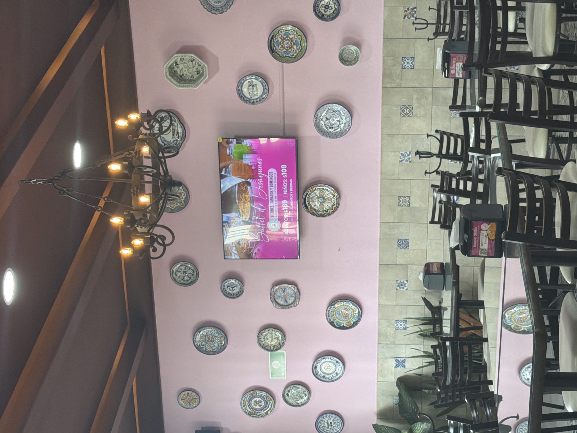 This media file depicts the interior of a restaurant, likely located in Puebla, Mexico, based on the architectural style and decor. A prominent feature is a light pink/purple wall, extensively decorated with numerous colorful and intricately patterned ceramic plates, reminiscent of Talavera pottery. A large, dark metal chandelier with multiple glowing bulbs hangs from a dark-beamed ceiling, casting warm light.
Centrally placed on the decorated wall is a TV screen displaying an advertisement for a "Buffet de Desayunos" (Breakfast Buffet). The ad specifies availability on "VIERNES, SABADOS Y DOMINGOS" (Fridays, Saturdays, and Sundays) starting at "8:30 AM" at the "SUCURSAL DAVIKLETA" (Davikleta Branch). Pricing is listed as "ADULTO $189" and "NIÑOS: $109," indicating costs in Mexican pesos, and includes "ALIMENTOS Y BEBIDAS" (Food and Drinks).
The floor is covered with light-colored tiles, interspersed with decorative blue and white patterned squares. Several dining tables with dark-colored chairs are arranged throughout the space; some chairs are fitted with white covers or cushions. On one table, a small sign is visible with the text "SABORCITO CORAZÓN" (Little Flavor Heart), accompanied by "Google" and "COMPARTE" (Share), likely encouraging customer engagement or reviews. No people are present, suggesting the image may have been captured before opening hours or during an off-peak time. The overall scene conveys a welcoming and traditionally decorated dining environment.