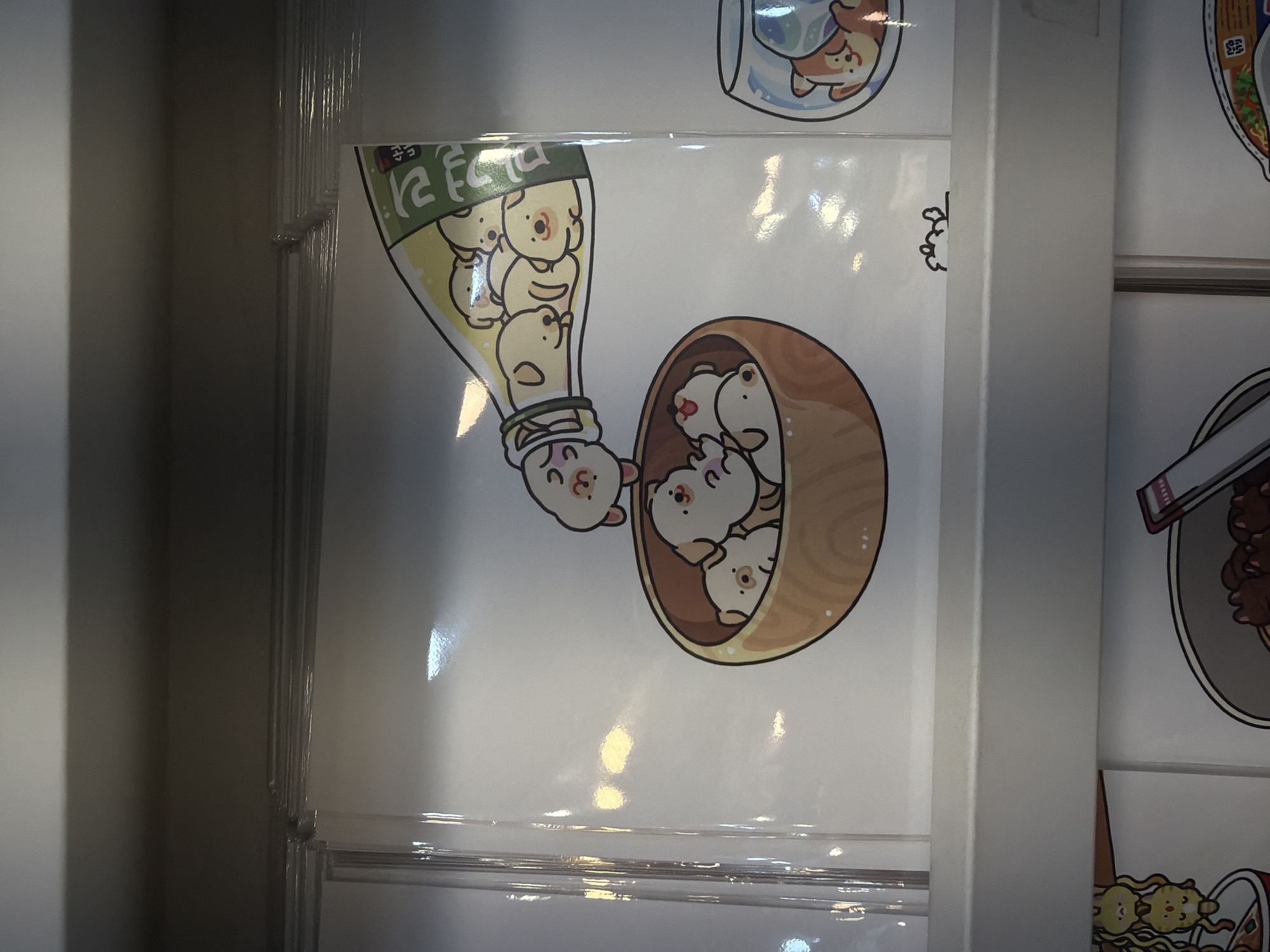 This image displays a section of a retail display, likely within a store in Seoul, South Korea, featuring an assortment of stickers or prints. The primary subject is a whimsical illustration of numerous small, light-colored dogs. One group of dogs is depicted spilling out of a green and white bottle, while another cluster is shown in a wooden bowl. A third illustration shows a dog in a fishbowl. The prints are encased in clear plastic sleeves and are arranged on shelves, suggesting they are merchandise for sale. The lighting in the scene is diffused, with some bright reflections indicating overhead lighting. The overall presentation is clean and organized, characteristic of a retail environment.