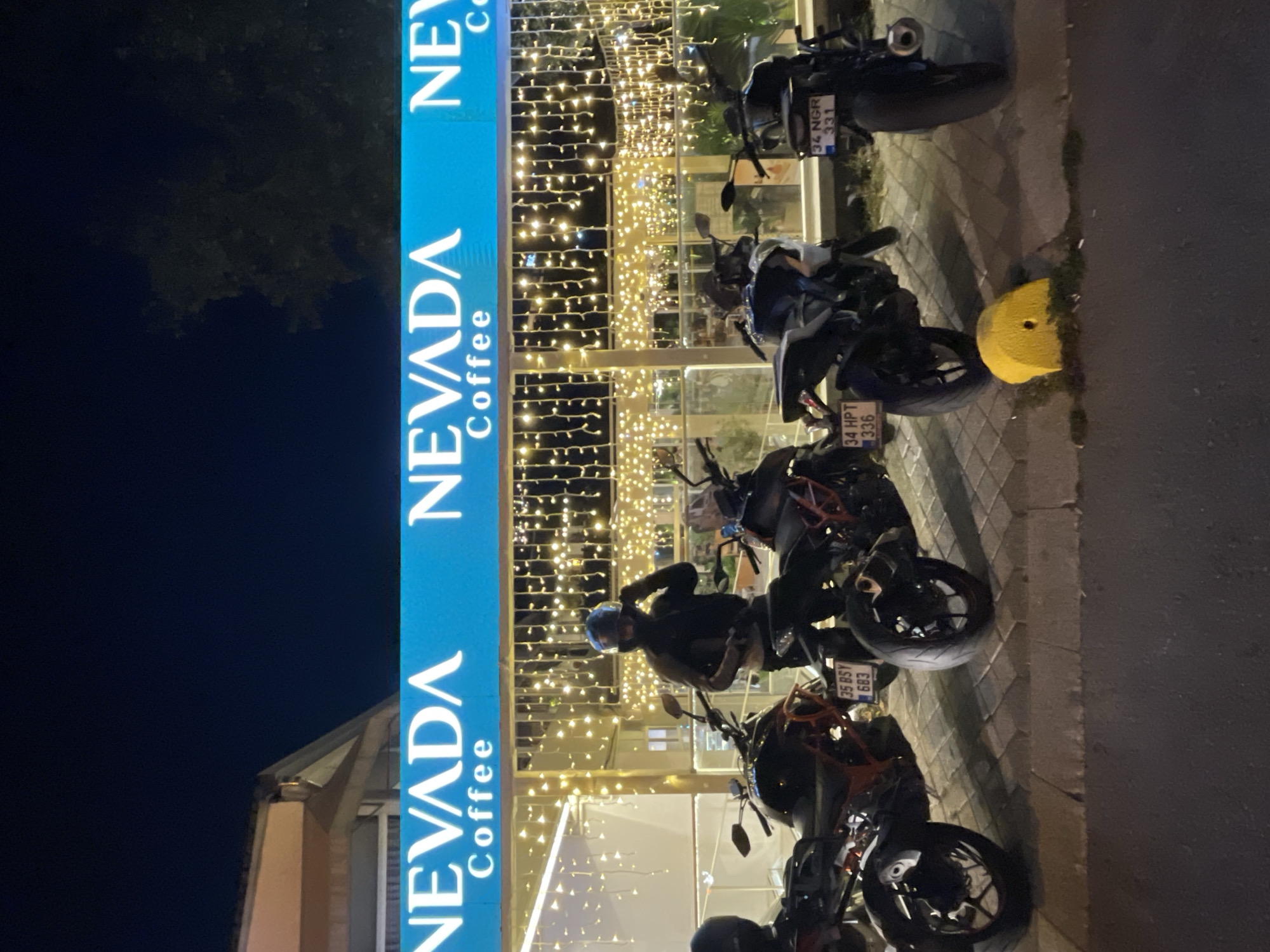 The image depicts the exterior of a "NEVADA COFFEE" establishment at night. The sign, illuminated in bright blue with white lettering, is prominent at the top of the frame. The building's facade is adorned with numerous strings of warm white fairy lights, creating a festive ambiance. In front of the establishment, parked along a sidewalk paved with rectangular bricks, are several motorcycles. One individual, wearing a helmet and dark clothing, is standing next to a motorcycle, appearing to adjust their helmet. The motorcycles are lined up, with license plates visible on some. The scene suggests a popular spot for motorcyclists, possibly a gathering point or a cafe frequented by the motorcycle community. The environment appears to be an urban setting, likely within Kadıköy, Turkey, given the presence of Turkish license plates.