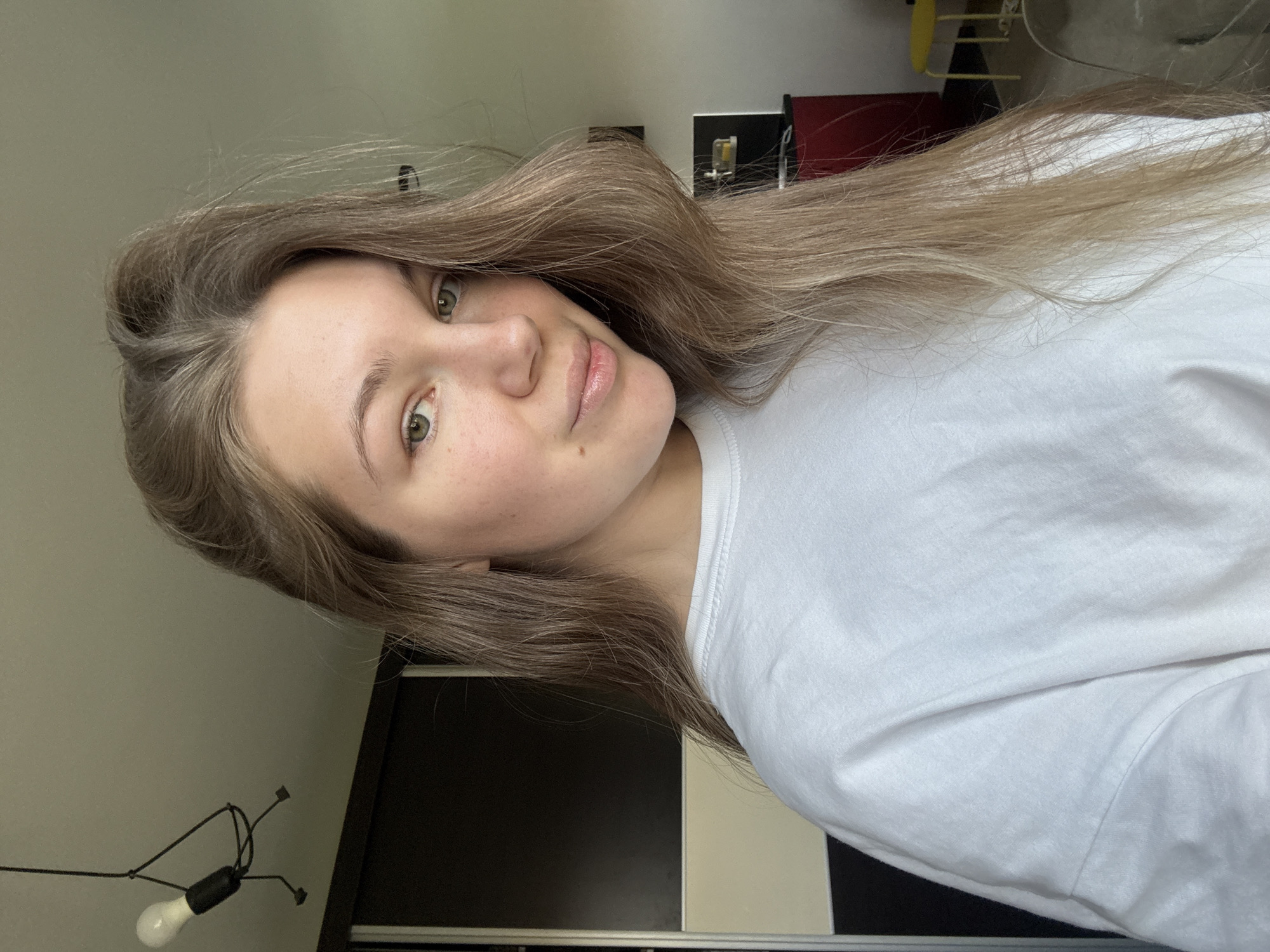 A young woman with long, light brown hair and green eyes is facing the camera. She is wearing a white t-shirt. Behind her is a wall with a modern light fixture and a closet or wardrobe. A glimpse of what appears to be a desk or table can be seen with items on it.
There is no indication in the image that the scene is located in Vozdovac Urban Municipality, Serbia.