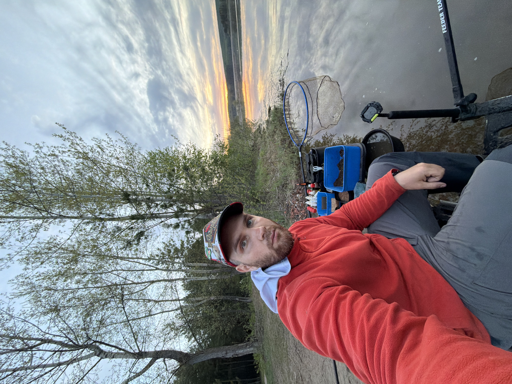 A man is seated in a chair, taking a selfie outdoors near a body of water. The man has a beard and is wearing a red fleece jacket, gray pants, a white undershirt, and a baseball cap. Behind him is a fishing net, a small blue bucket, and fishing equipment. In the background, there are trees and the body of water, with a colorful sky suggesting either sunrise or sunset. Based on "FEEDER TEAM" on the fishing rod, the person appears to be a feeder fisherman. While location details cannot be determined with absolute certainty from the image, the request identifies the location as Chaikovsky, Russia.