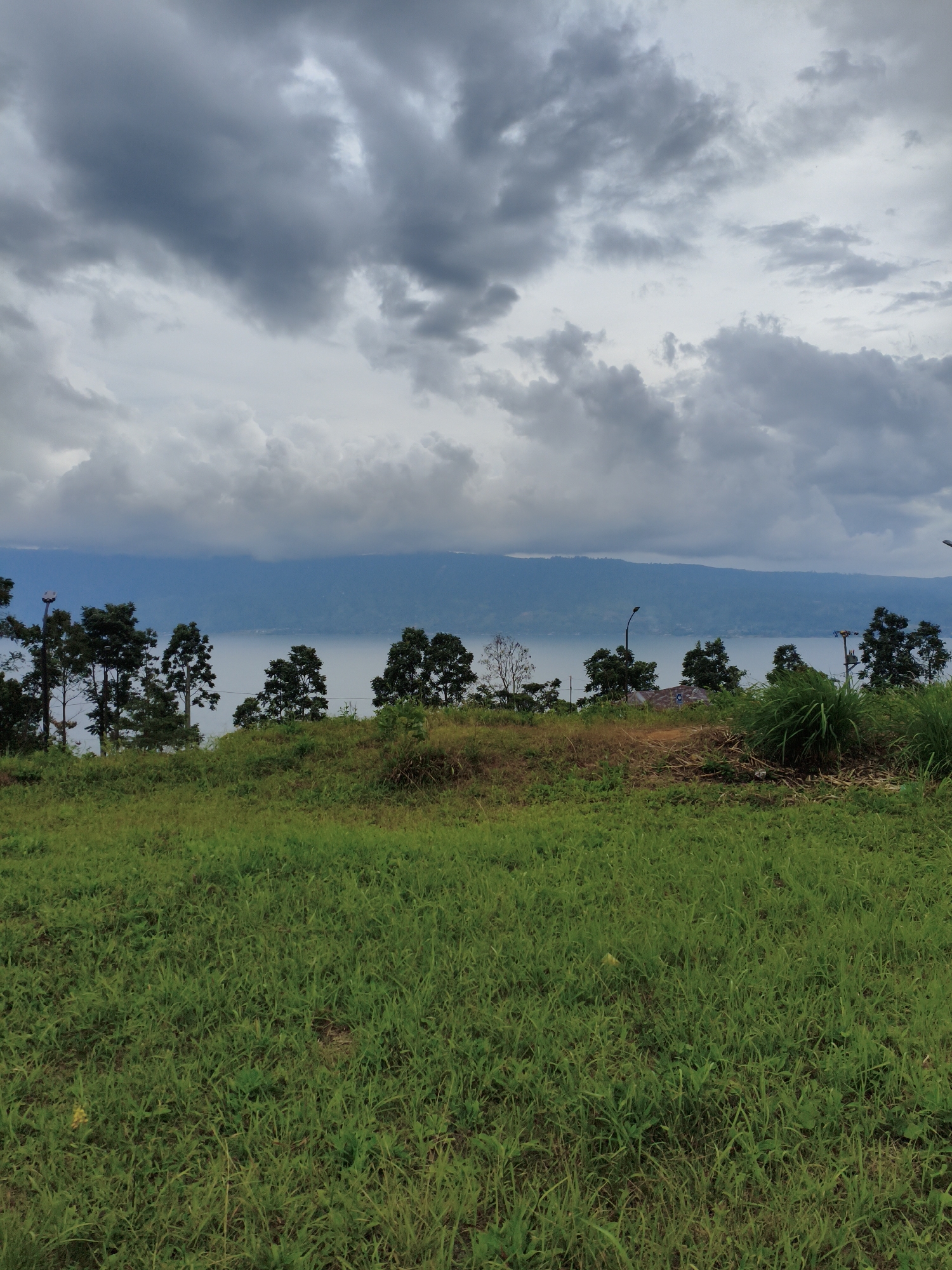The image depicts a serene natural landscape, likely an elevated viewpoint overlooking a body of water. In the foreground, a lush green grassy field slopes gently upwards, with patches of dry grass and soil visible in some areas. The middle ground is characterized by a line of trees, predominantly conifers with a few deciduous trees interspersed, standing on the edge of the terrain. Beyond the trees, a vast, calm body of water stretches horizontally, reflecting the overcast sky. In the distance, a range of hills or mountains forms the horizon, their outlines softened by atmospheric haze. The sky is filled with dramatic, dark gray clouds, suggesting an impending storm or recent rainfall. There are no visible people, structures, or specific landmarks that would indicate the location within Pem.tambun Raya, Indonesia. The scene is tranquil and dominated by natural elements.
