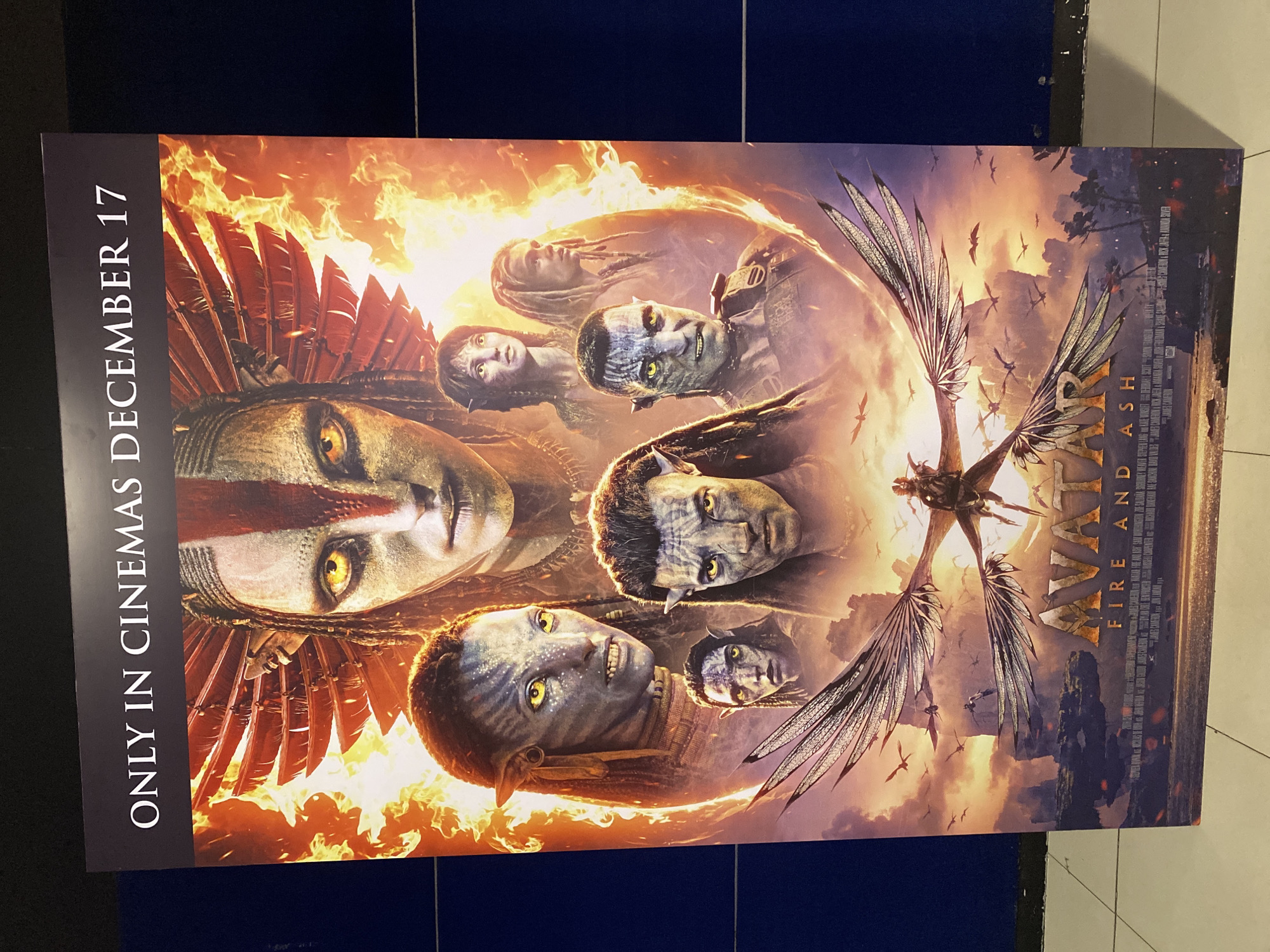 This is a vertically oriented movie poster for "Avatar: Fire and Ash," displayed indoors, likely in a cinema lobby in Davao City, Philippines. The poster features a dramatic, action-oriented scene with a fiery and smoky background, suggesting intense conflict.
The main subjects are several Na'vi characters, depicted with blue/grey skin and striking yellow eyes. A large Na'vi figure dominates the left side, wearing a prominent red-feathered headdress. Other Na'vi faces are visible throughout the poster, some with intricate facial patterns or determined expressions. One character in the upper right appears to be wearing some form of military-style gear. In the center-right, a figure is shown riding a large winged creature, possibly an Ikran or Banshee, with intricate, almost insect-like wings. Silhouettes of other flying creatures are scattered across the sky.
Visible text includes "ONLY IN CINEMAS DECEMBER 17" running vertically along the left edge. The movie title "AVATAR FIRE AND ASH" is prominently displayed on the right side, with the "Avatar" logo in a metallic, stylized font and "Fire And Ash" in smaller, fiery letters below. A block of smaller white text, likely movie credits, is also visible on the right. The overall atmosphere conveyed is one of epic adventure, fantasy, and impending conflict.
