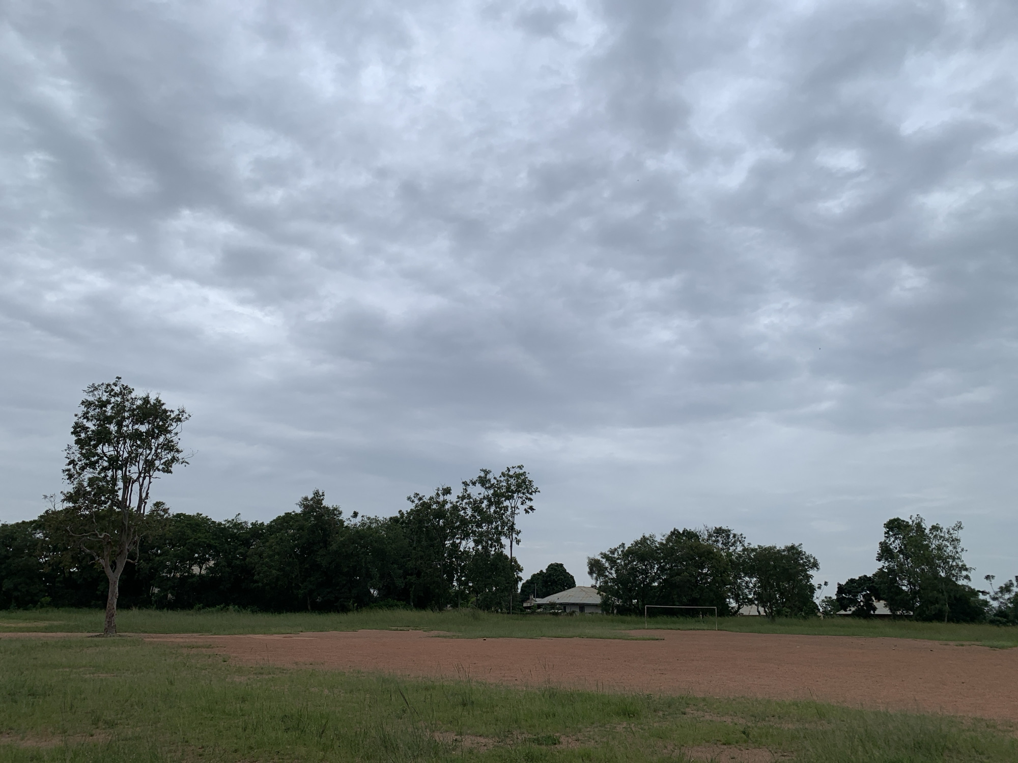 The image depicts an outdoor scene in Kuru Karama, Nigeria, under an overcast sky. In the foreground, there is a dusty, reddish-brown field, possibly a sports field, with patches of green grass. Several trees are visible, with one particularly tall, slender tree standing to the left of the frame. Behind the field, a line of dense, dark green trees forms a natural backdrop, with some buildings and a white soccer goal visible among them. The sky is filled with gray clouds, suggesting an impending rain or a generally cloudy day. There are no people or animals visible in the image.