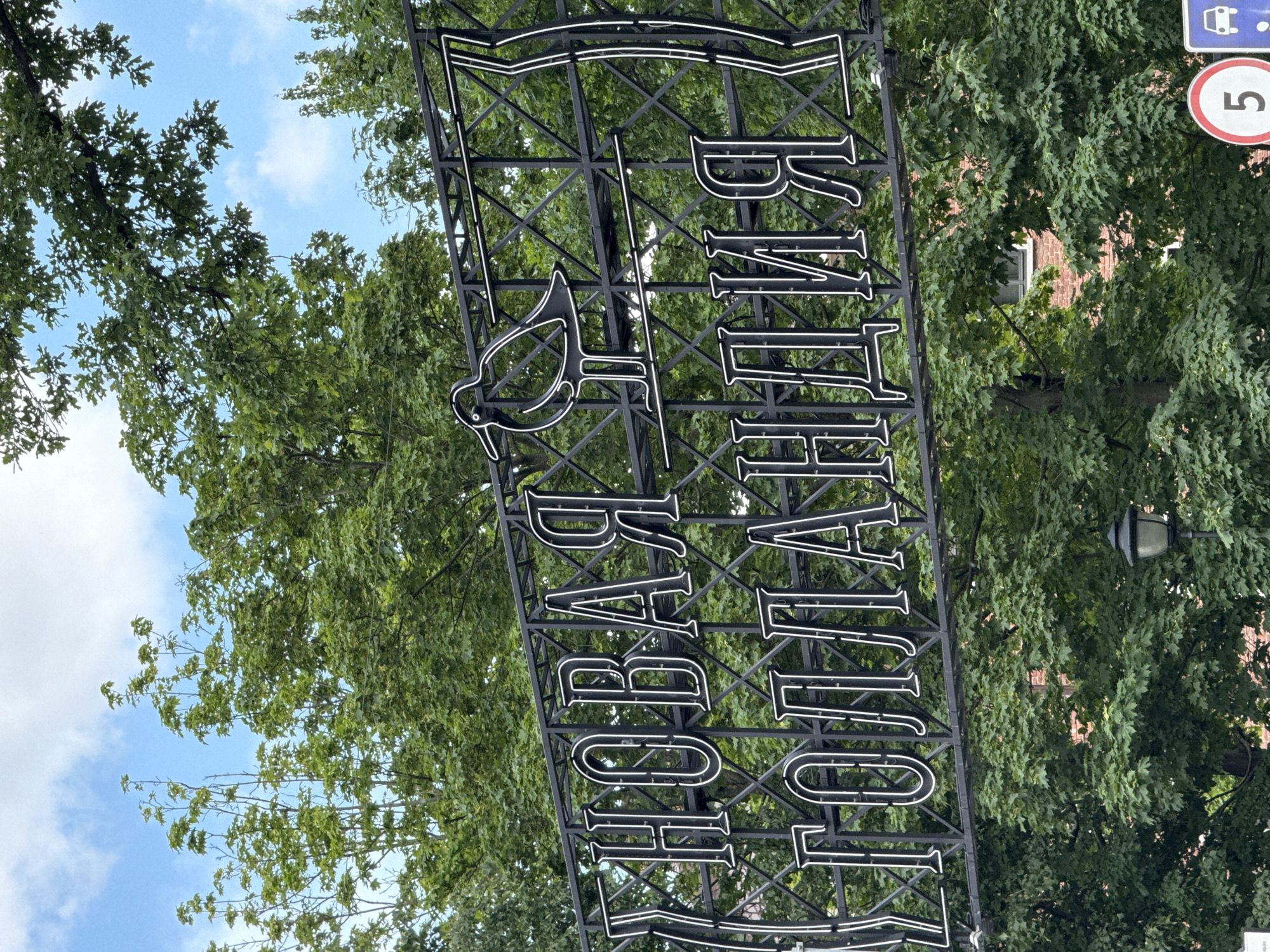 The image shows a neon sign in Cyrillic script reading "НОВАЯ ГОЛЛАНДИЯ" (New Holland) in front of lush green trees and a blue sky with scattered clouds. The sign is black with white neon outlines for the letters and features a stylized bird silhouette. The trees are dense and leafy, with some brick buildings visible in the background. In the lower right corner, there are traffic signs, including a white circle with a red border and the number "5" inside, and a blue rectangular sign with a white car icon. A lamppost is also visible behind the sign. The scene appears to be an outdoor public space in Saint Petersburg, Russia, likely an entrance to the New Holland Island cultural space.