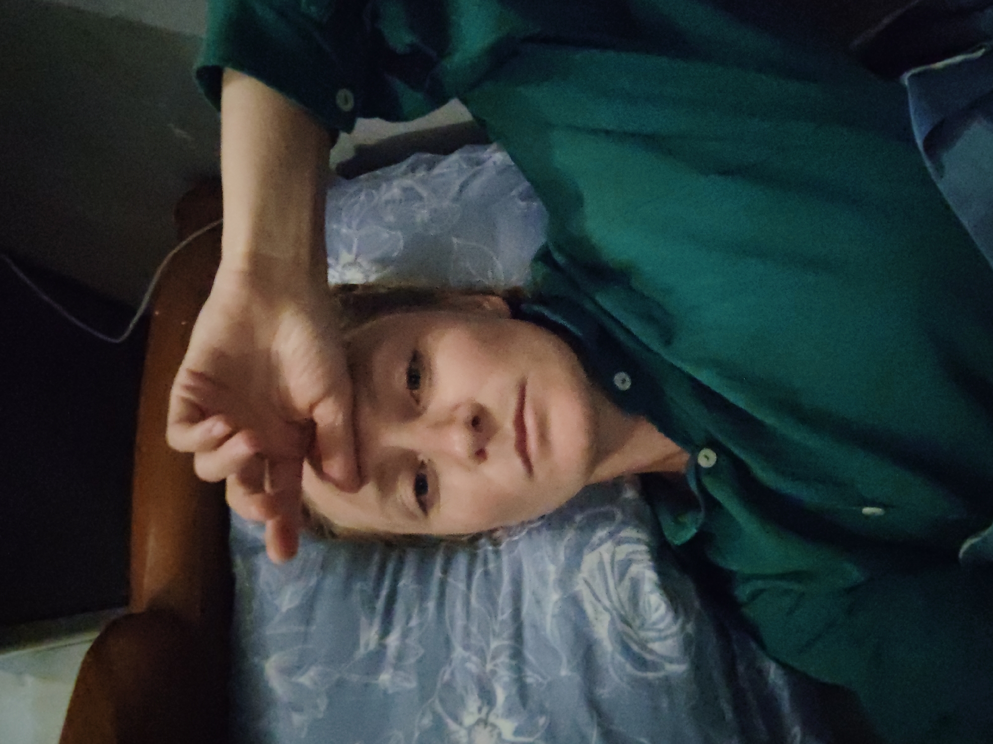 A fair-skinned individual is lying on a blue and white floral patterned pillow and possibly a bed, wearing a green long-sleeved collared shirt. The individual's head is tilted to the left, and their left hand is raised, obscuring the left side of their face. Part of a dark wooden bed frame is visible in the background. The setting appears to be indoors. Location data not included.