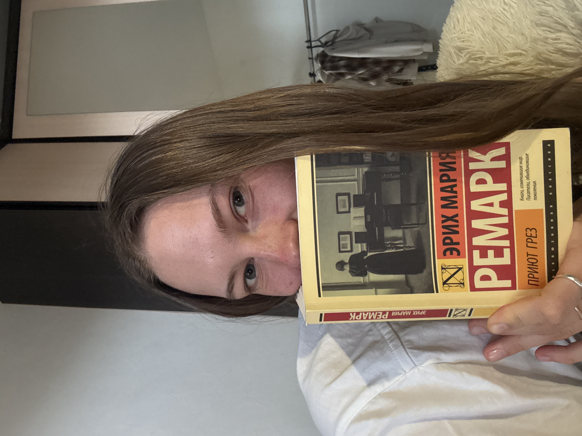 A close-up portrait shows a young person, seemingly female, with long brown hair, holding a book in front of their face. Their right eye and forehead are visible above the book, looking directly at the camera with a neutral expression. They are wearing a light-colored, possibly white, collared shirt.
The book has a yellow and red cover with Russian text. The prominent author's name reads "ЭРИХ МАРИЯ РЕМАРК" (Erich Maria Remarque), and the title is "ПРИЮТ ГРЕЗ" (Priut Grez), which translates to "The Dream Room" or "The Dream Refuge." A small emblem is situated between the author's first and last names. Additional text on the cover includes "Книги, изменившие мир. Писатели, объединившие поколения." (Books that changed the world. Writers who united generations.) and "Эксклюзивная классика" (Exclusive Classics). The book's cover art features a black and white image of a person's silhouette seated at a table in a room with framed pictures on the wall.
In the background, to the right of the person's head, there is a fluffy white or cream-colored object, likely a blanket or pillow, and a glimpse of a closet with clothes on hangers. The setting appears to be an indoor, casual environment. The overall mood is calm and engaging. The text on the book is in Russian. This scene is contextualized as being in Perm, Russia.