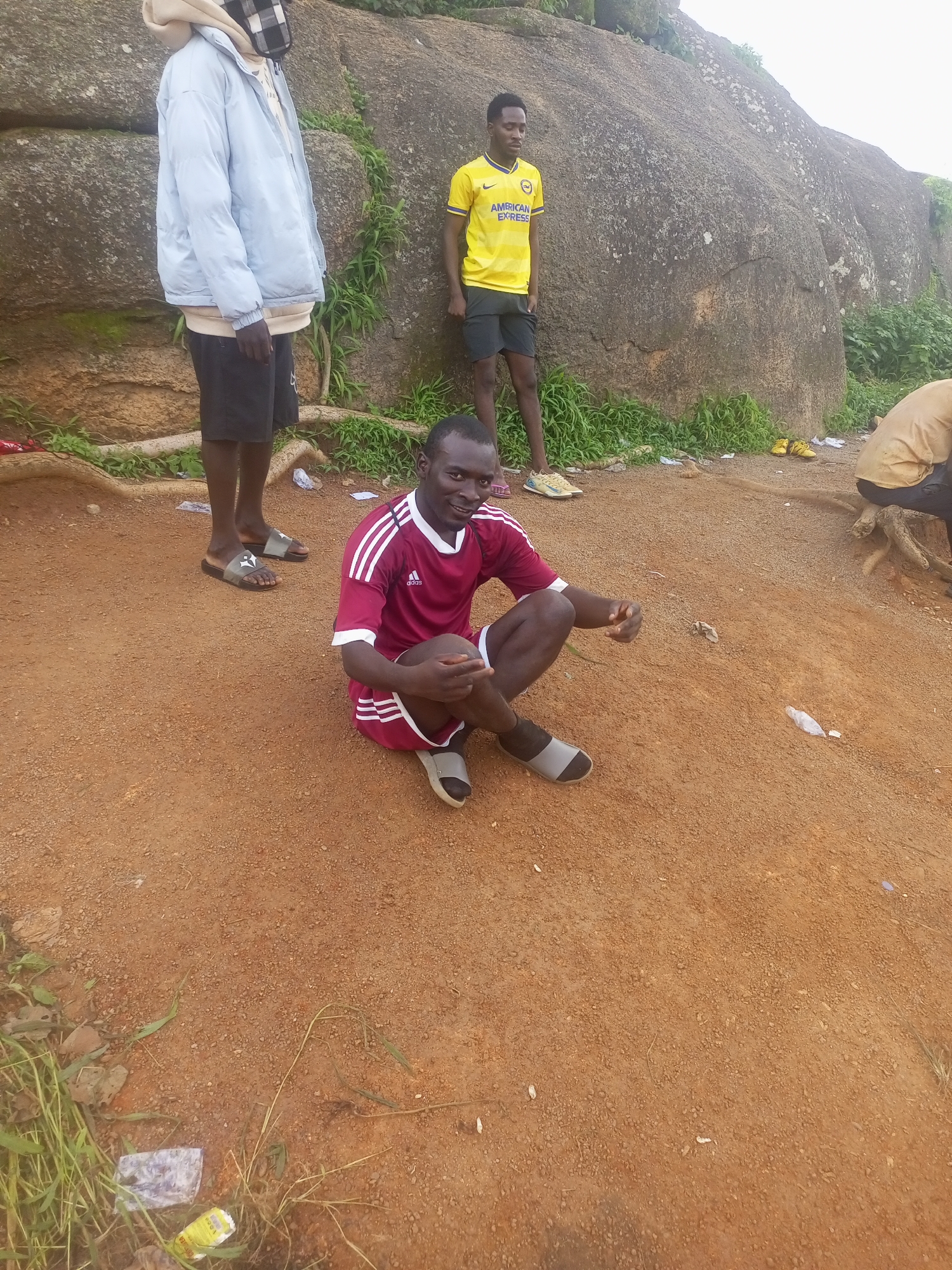 In the city of Jos, Nigeria, a group of young men are gathered near a large rock formation. The ground is covered in reddish-brown soil with scattered pebbles and dry grass. To the left, one man stands wearing a light blue puffer jacket over a tan hoodie and dark shorts, his face obscured by a checkered mask. Beside him, another man stands in a yellow and white striped t-shirt with "AMERICAN EXPRESS" printed on it, wearing dark shorts and light-colored athletic shoes. He is looking down. In the foreground, a man in a maroon athletic jersey with white stripes down the sleeves and white shorts sits on the ground, smiling at the camera. He is wearing grey and black slide sandals. To the right, another person, partially visible, is seated with their back to the camera, wearing a tan shirt and dark pants. There is scattered litter on the ground, including plastic wrappers and bottles. The overall impression is a casual gathering outdoors.