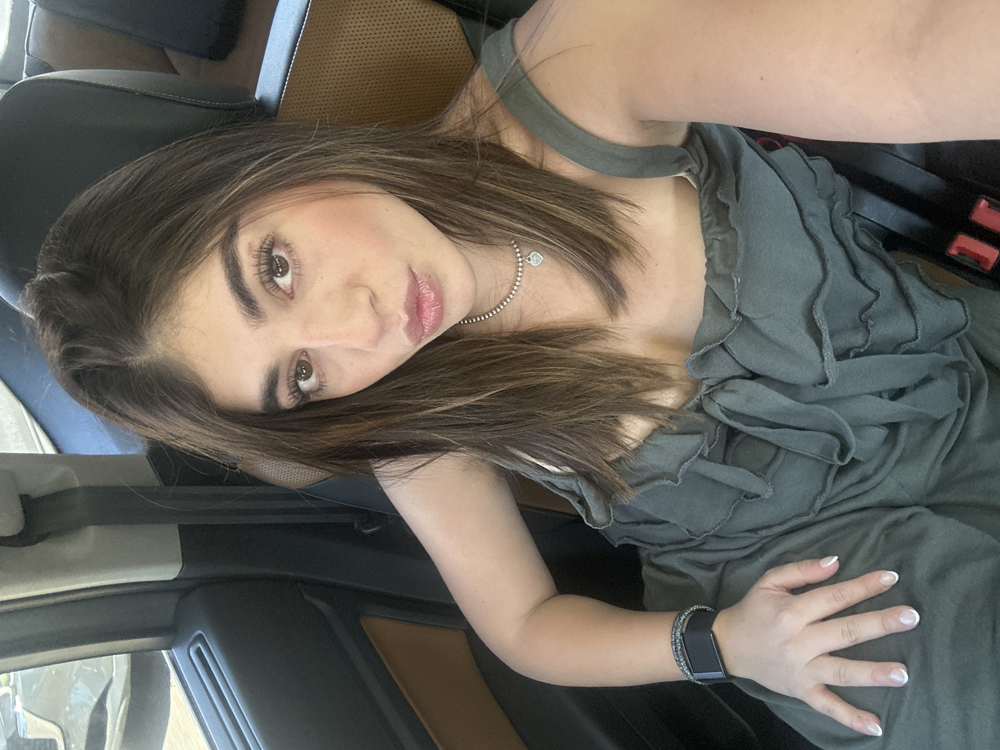 A woman is reclining in what appears to be a car. She has brown hair with blonde highlights, brown eyes, and light skin. She is wearing a grey top with ruffled detailing and a silver necklace. On her left wrist, she wears a black smartwatch. The car's interior is tan and black. The setting appears to be daytime inside the car. No specific details indicate the location in Mexico City, Mexico.