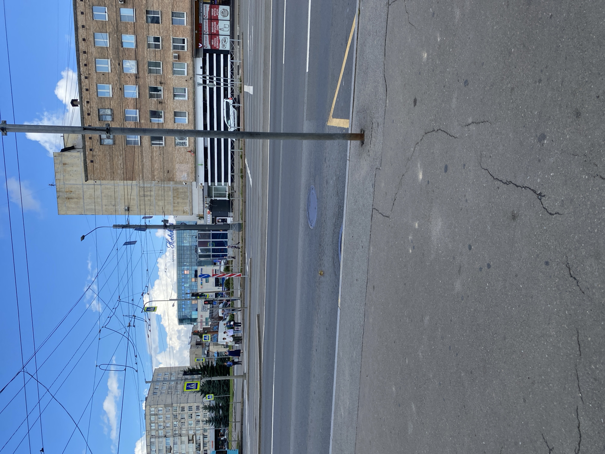 The image depicts a street scene in Saint Petersburg, Russia, on a bright, cloudy day. In the foreground, a grey asphalt sidewalk with cracks is visible, leading to a street with lane markings and a yellow bus stop marker. Across the street, several people are standing at a crosswalk. Behind them, a modern glass-fronted building with the word "Heboka" is prominent, alongside other commercial establishments with signs in Russian. Further in the background, a taller, older brick building with numerous windows and a lighter colored concrete section on top dominates the skyline, with a multi-story apartment building to the left. Overhead, a complex network of trolleybus wires crisscrosses the sky, with a grey metal pole in the center of the frame. The overall environment suggests an urban setting with public transportation infrastructure and commercial activity.