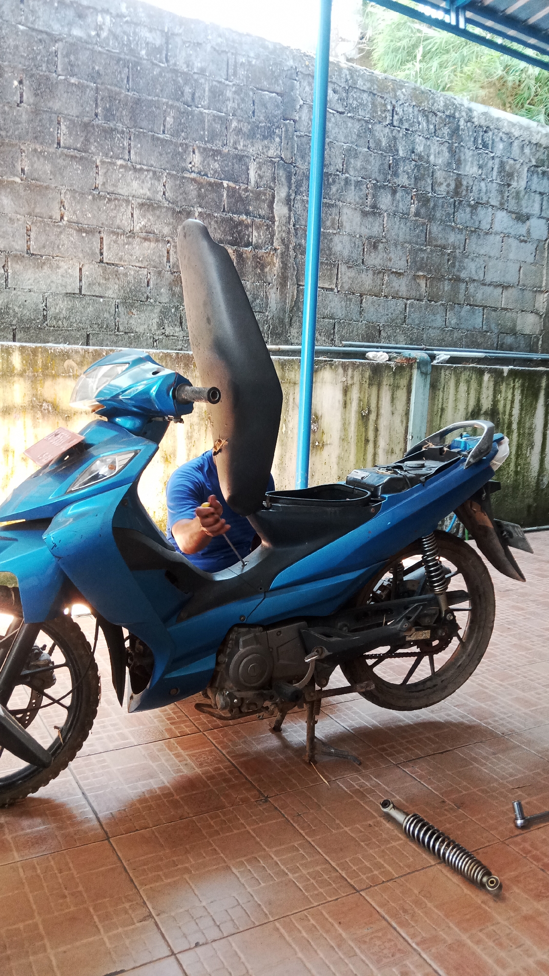 A person in a blue t-shirt is working on a blue and black motorcycle. The motorcycle seat is propped open, revealing the undercarriage. The person is holding a tool, possibly a screwdriver, and appears to be working on a screw or bolt. The motorcycle is parked on a tiled floor. In the background, there is a grey brick wall and a blue pole. The overall scene suggests a repair or maintenance activity taking place outdoors, possibly in a residential area of Jawa, Indonesia.