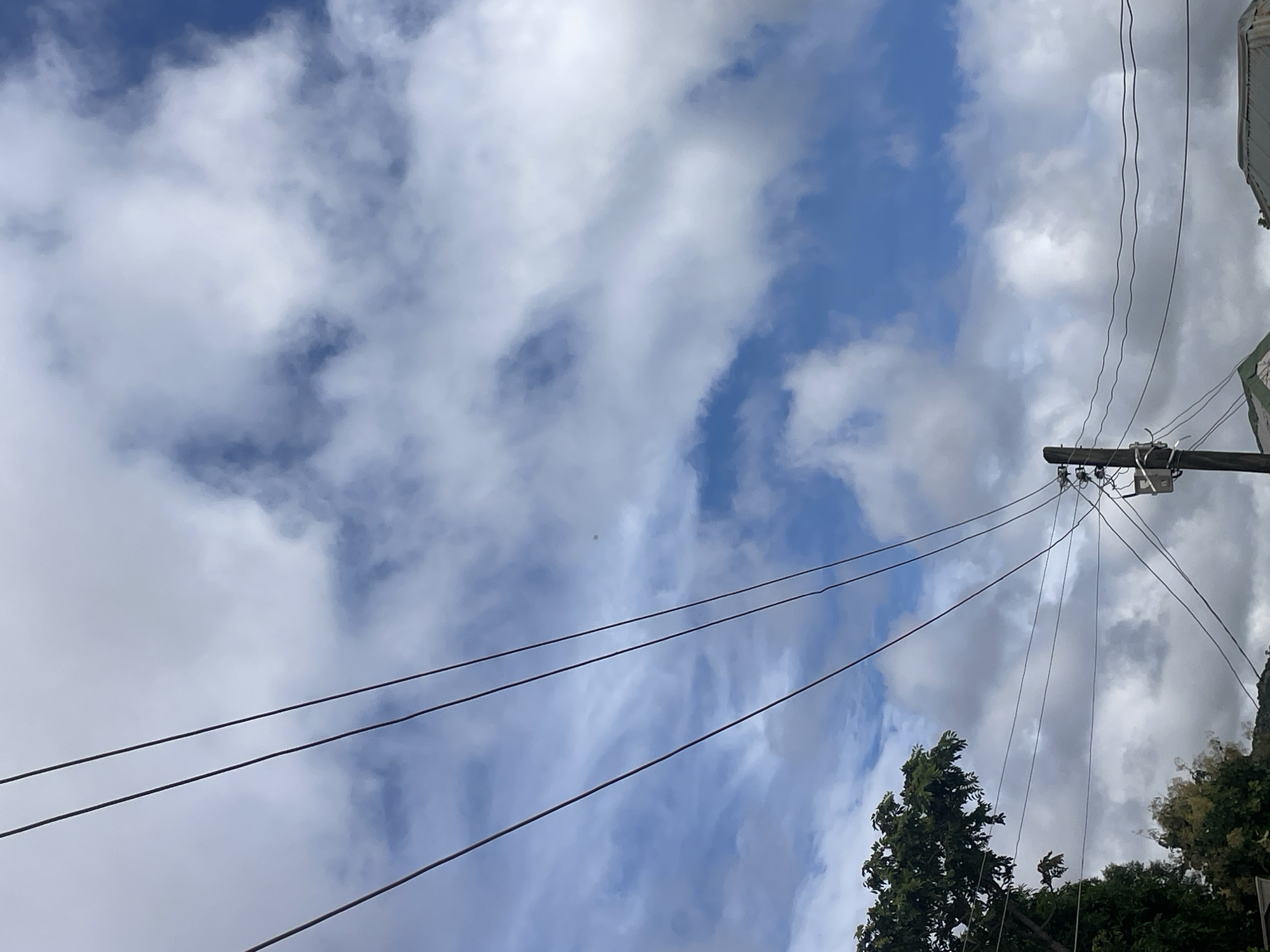 The image depicts a view of the sky with a cluster of clouds against a blue background. Several power lines stretch across the frame, running diagonally from the upper left to the lower right. A utility pole with electrical equipment is visible on the right side of the image, supporting some of these lines. In the lower left corner, the top of a large green tree is visible, with its branches reaching upwards. Below the tree and further into the background, a hint of smaller trees and foliage can be seen. The sky suggests it is daytime, possibly with some overcast conditions due to the density of the clouds. The scene appears to be an outdoor environment within a residential or semi-urban area, consistent with a location in Jos, Nigeria. No people or specific actions are identifiable in the image.