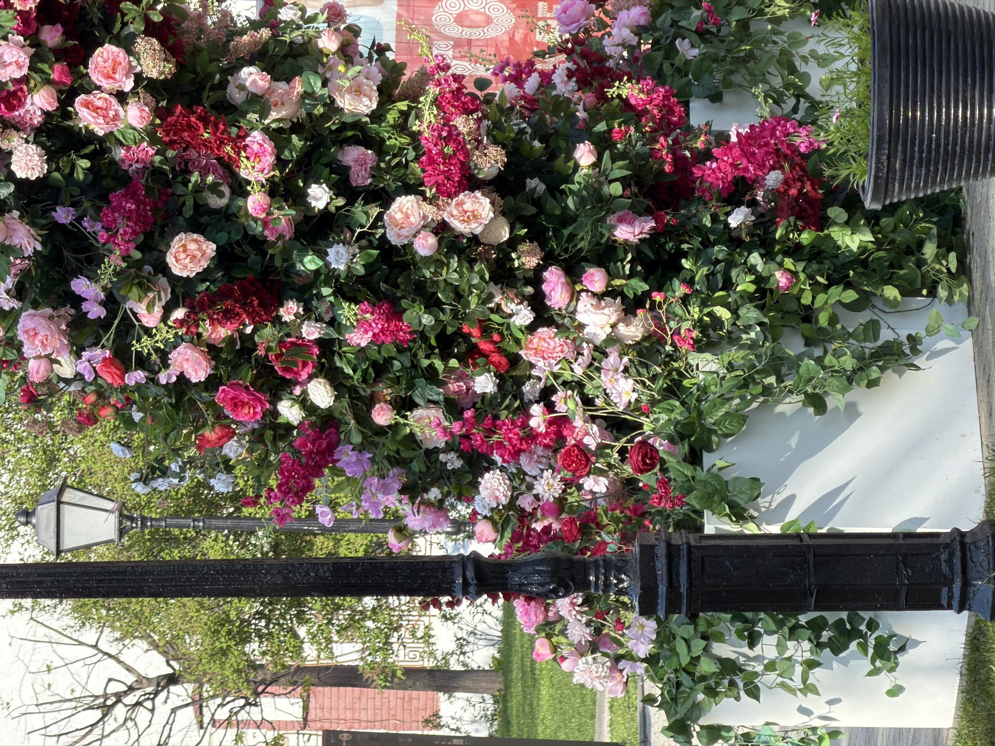 A vivid floral display adorns a location in Moscow, Russia. The arrangement showcases a variety of pink and red flowers amidst lush green foliage. A black lamppost is present, further entwined with the floral elements. The backdrop consists of a white wall and glimpses of greenery. The scene suggests an outdoor setting within the urban environment of Moscow.