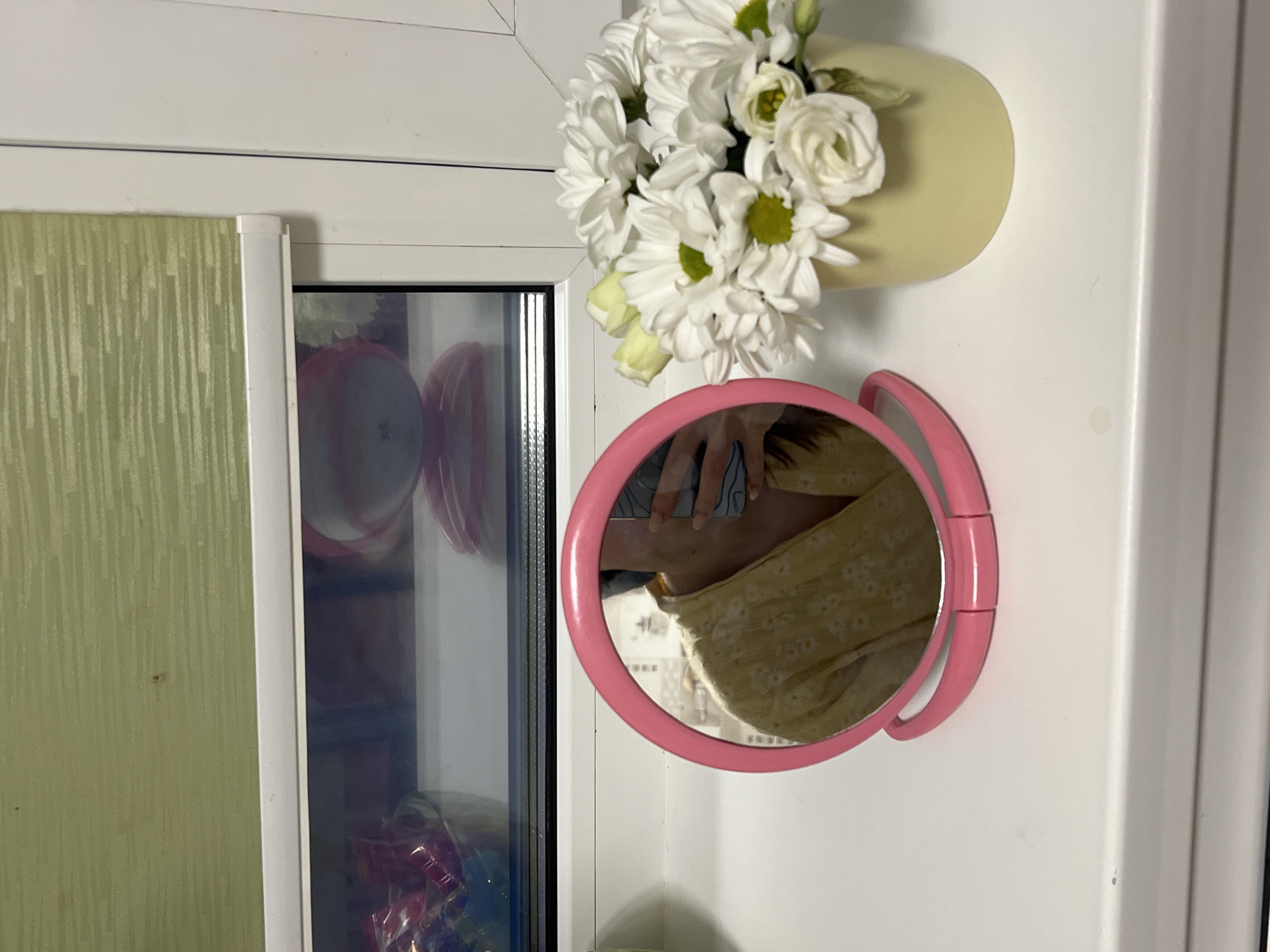 A close-up shot indoors features a portion of a window frame on the left, with a pale green textured wall visible through the open window. Adjacent to the window, on a white surface, is a light yellow vase holding a bouquet of white flowers, including daisies and roses. Below the flowers, a round, pink mirror reflects a person wearing a yellow floral shirt. The environment appears to be domestic, and the composition suggests an interior setting.