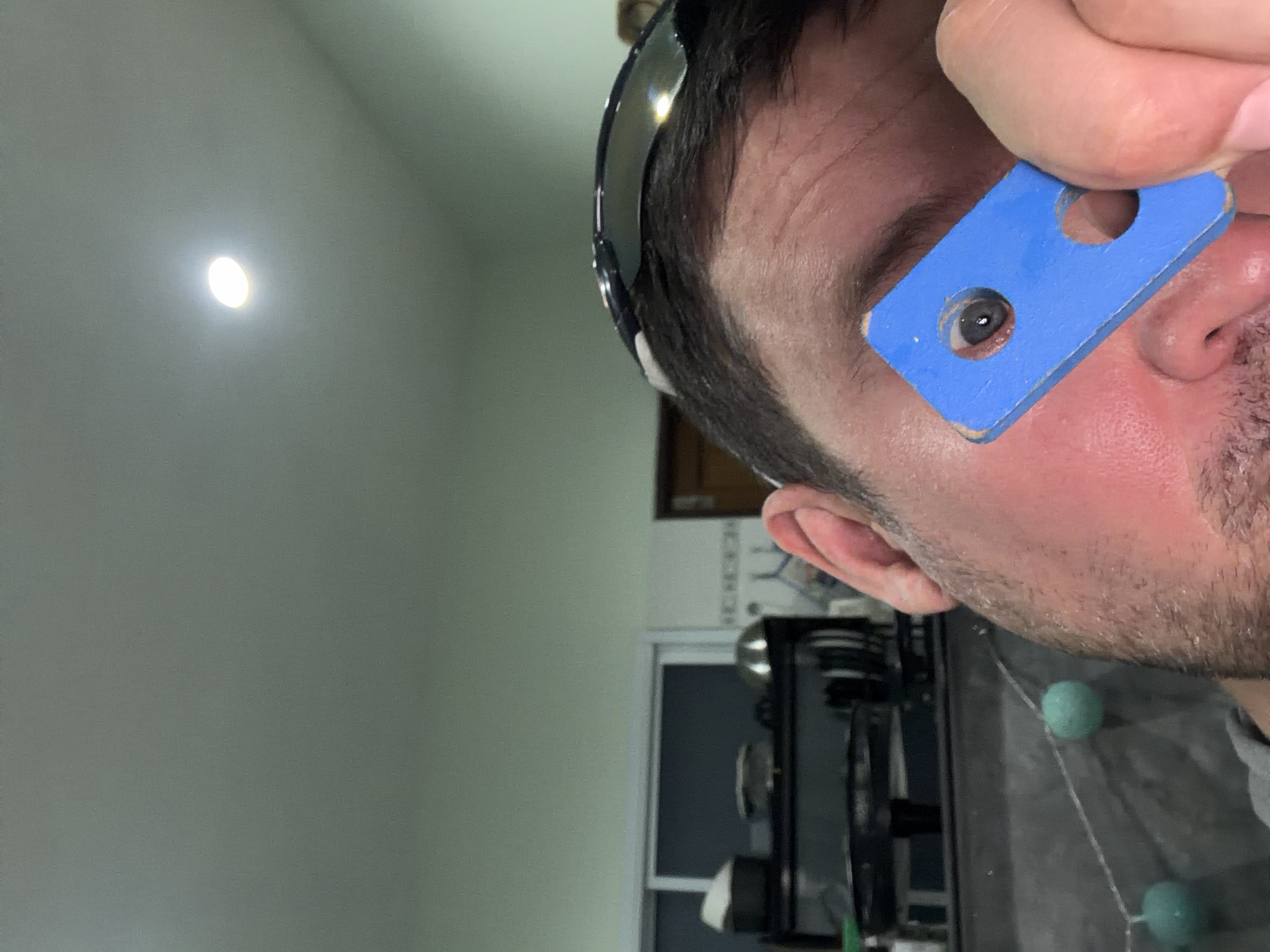 In Baan Mae Nam, Thailand, a close-up, eye-level, indoor shot shows a person's face partially obscured by a blue rectangular object with two circular cutouts. The person is wearing black-rimmed sunglasses. One eye is visible through the larger cutout, revealing a dark iris. The background suggests a domestic setting, with a ceiling light visible at the top and kitchen appliances and cabinetry in the blurred background. The lighting is dim, with the ceiling light providing the primary illumination.