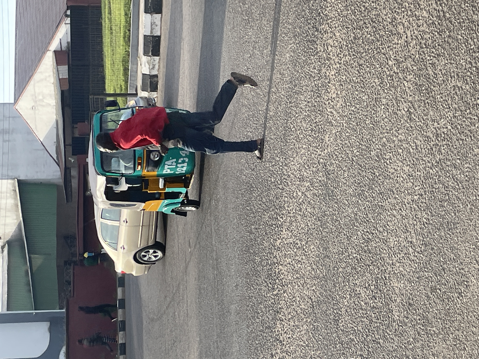 In Jos, Nigeria, a man in a red shirt and dark pants is captured in motion, running towards the right side of the frame. He is positioned in front of a turquoise and yellow motorized tricycle, commonly known as an "Okada." The tricycle bears the registration number "PTA 12134" in white lettering on its side. Behind the tricycle, a light-colored sedan is visible, parked or moving along the road. The scene unfolds on a paved road with a checkered black and white curb bordering the left side. In the background, a building with a white, triangular roof stands prominently, with a green structure to its left. A patch of green grass is also visible, suggesting an outdoor, urban setting. The overall atmosphere is one of everyday street activity in a Nigerian city.