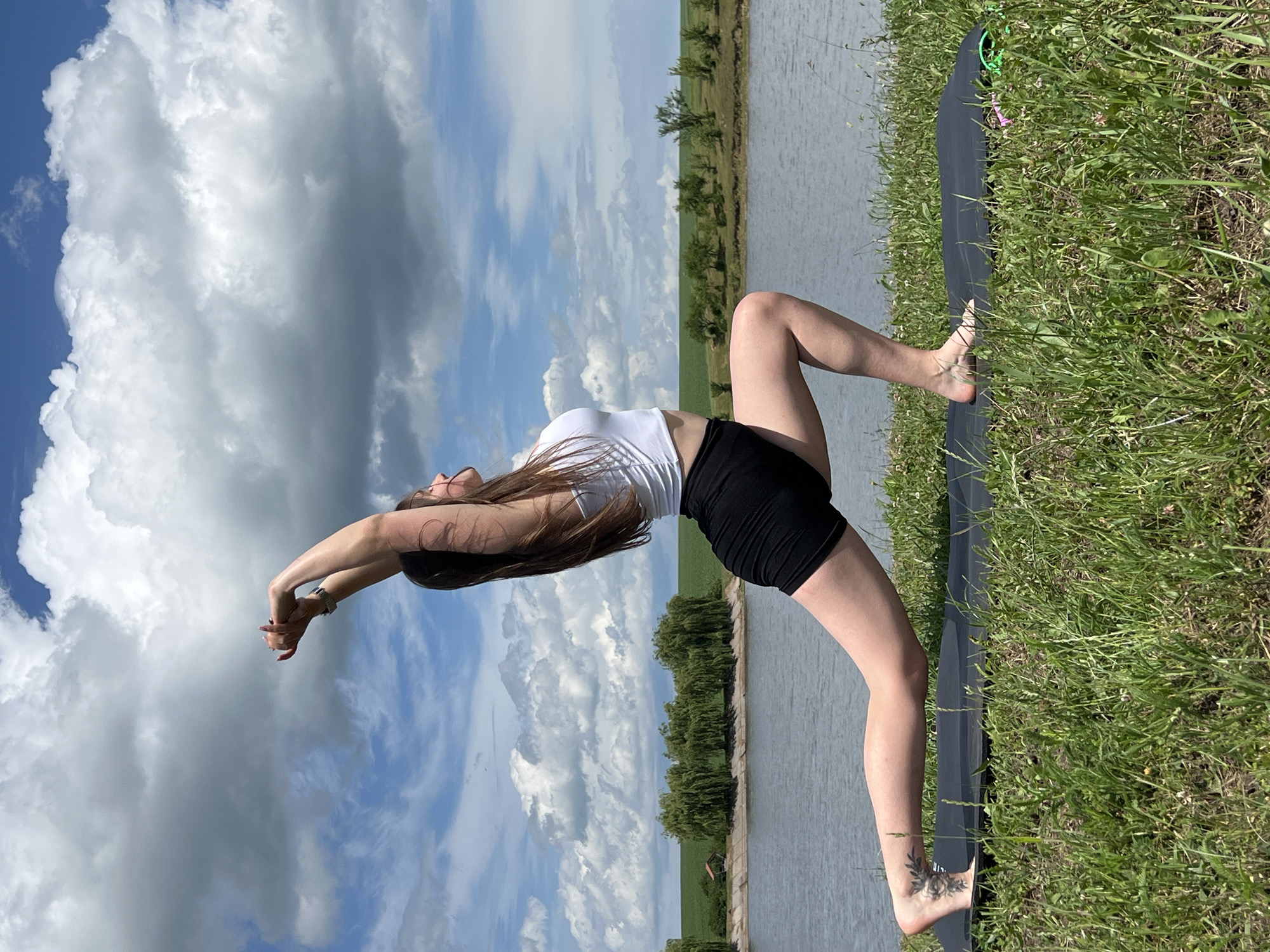 A young woman with long brown hair is performing a yoga pose outdoors on a mat. She is wearing a white tank top and black shorts. Her arms are extended above her head, and her body is positioned in a lunge. The background features a calm body of water, lush green grass, and a cloudy sky. The scene appears to be set in a natural landscape near the village of Borychivka, Ukraine.
