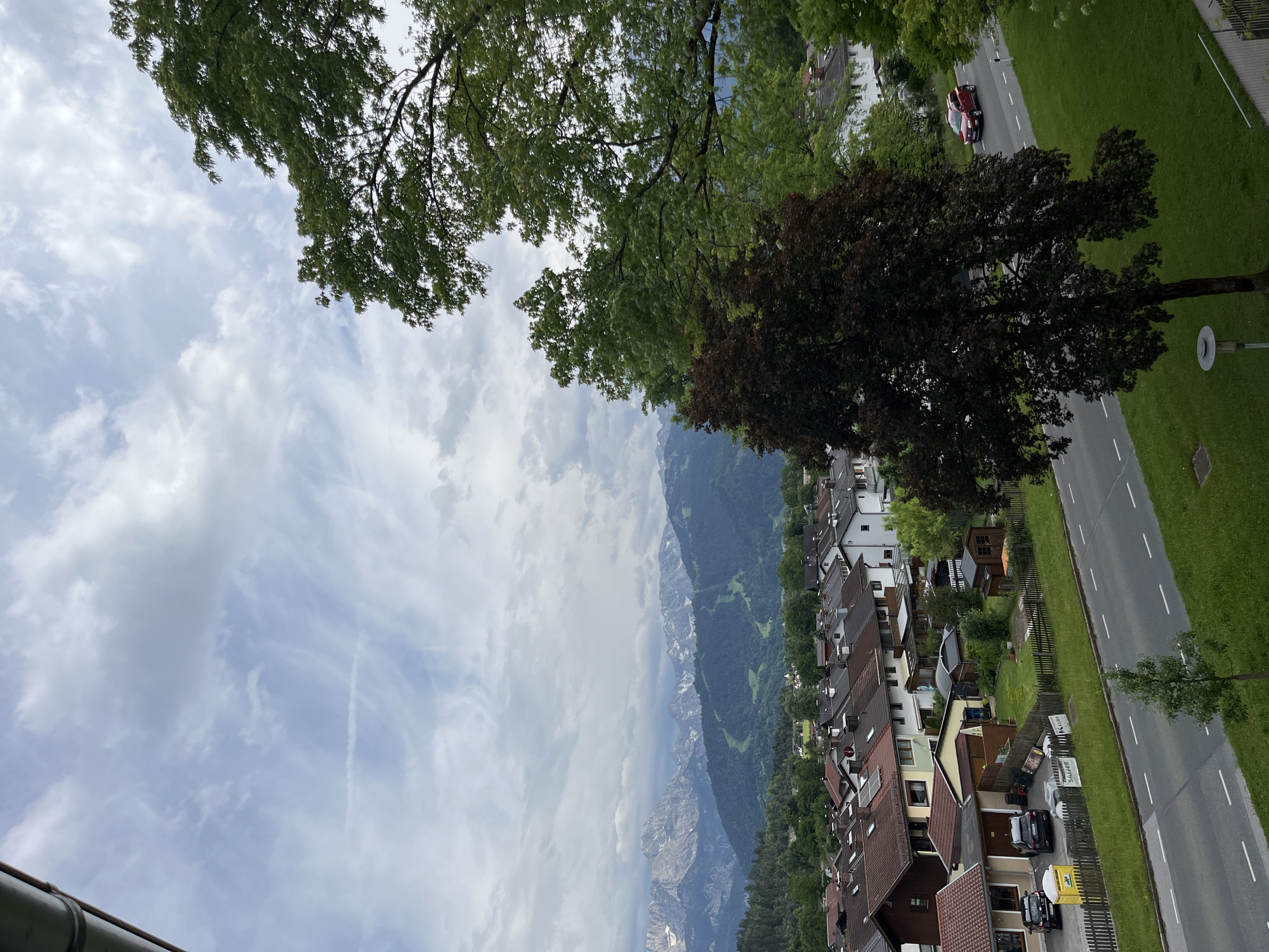 The image depicts a scenic view of Garmisch-Partenkirchen, Germany. The scene is dominated by the dramatic backdrop of snow-capped mountains, partially obscured by clouds. In the foreground, a quaint German village stretches along a paved road. The houses are characterized by their traditional architecture, featuring red-tiled roofs and white or light-colored facades.
The time of day appears to be overcast, with a bright, cloudy sky suggesting daytime, likely mid-morning or afternoon. The weather seems mild, as indicated by the lush green foliage of trees and well-maintained lawns. A dark red car is visible on the road, adding a touch of color to the scene.
Notable details include the dense treeline bordering the village, the presence of fences separating properties, and what appear to be solar panels on some rooftops. A few signs are visible, one of which has text in German, but it is too small to be legible. The overall impression is one of a peaceful, picturesque mountain town on a cloudy day. No people are explicitly visible in the image.