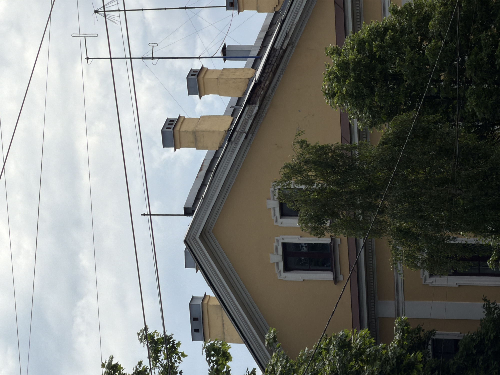 The image, originally oriented vertically and rotated clockwise, depicts the upper portion of a building and its surroundings under an overcast sky. The building features a pale yellow facade and a steep, grey-colored roof. Multiple prominent chimneys, square in shape and painted yellow with grey caps and small openings, are visible along the rooftop.
Several windows with dark frames and decorative white or light grey surrounds are set into the building's facade. Green foliage from trees partially obscures parts of the building at the bottom and right edges of the frame.
Above the building, against a bright but cloudy sky, a network of overhead power lines is visible, along with at least two distinct antennas. One antenna appears to be a multi-element array, while another is simpler and T-shaped. The soft, diffuse lighting indicates it is daytime. No people or visible text are present in the image. The architectural style suggests an urban setting, consistent with its given location in Saint Petersburg, Russia.