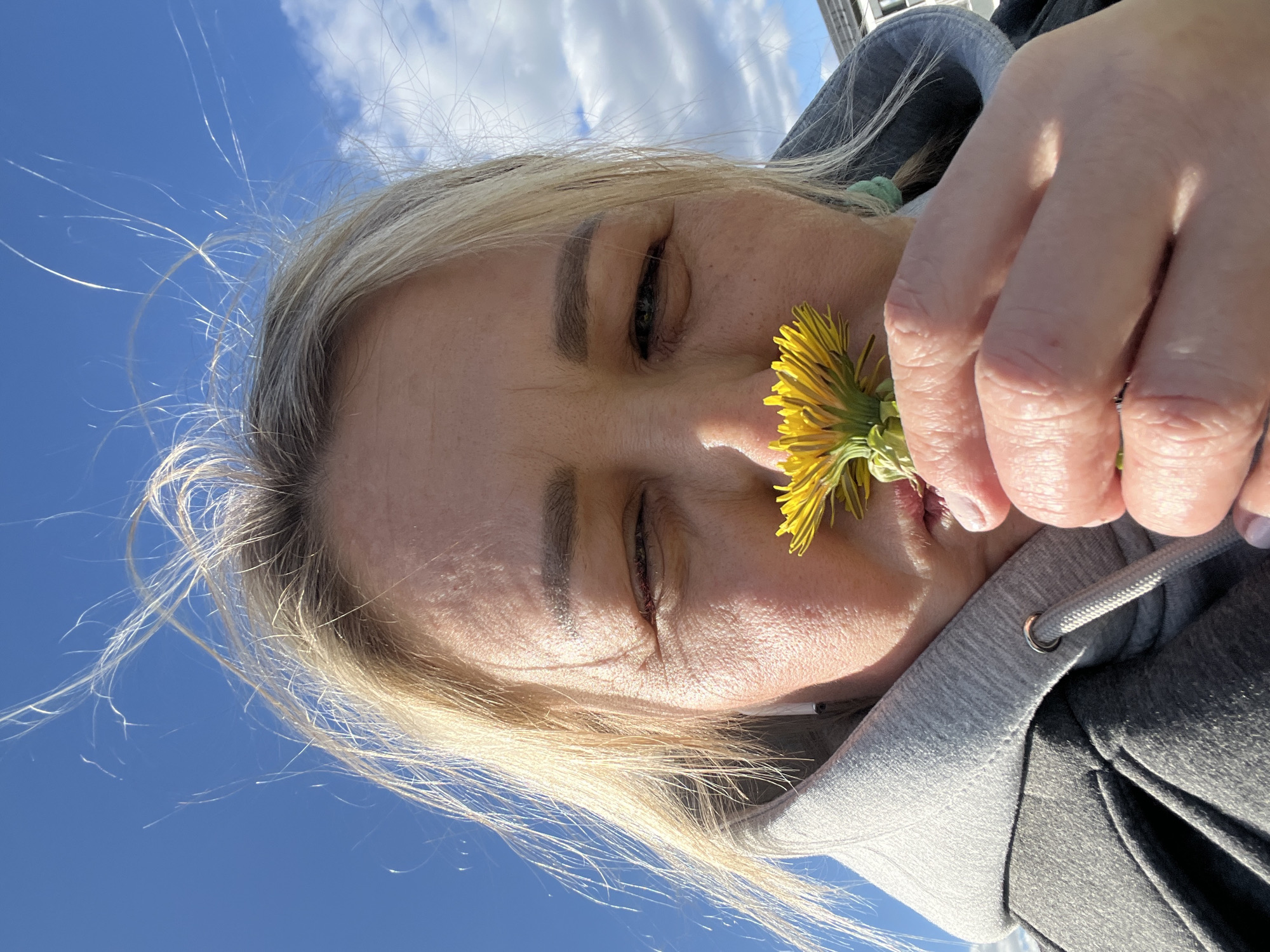 The image shows a woman holding a yellow dandelion flower up to her face. The background is a bright blue sky with some white clouds. The woman has blond hair and is wearing a gray hooded sweatshirt. The photo appears to be taken outdoors, possibly in a park or natural area. The image does not contain any information or features that allow for identification of the location as Chaikovsky, Russia, or any specific city for that matter.