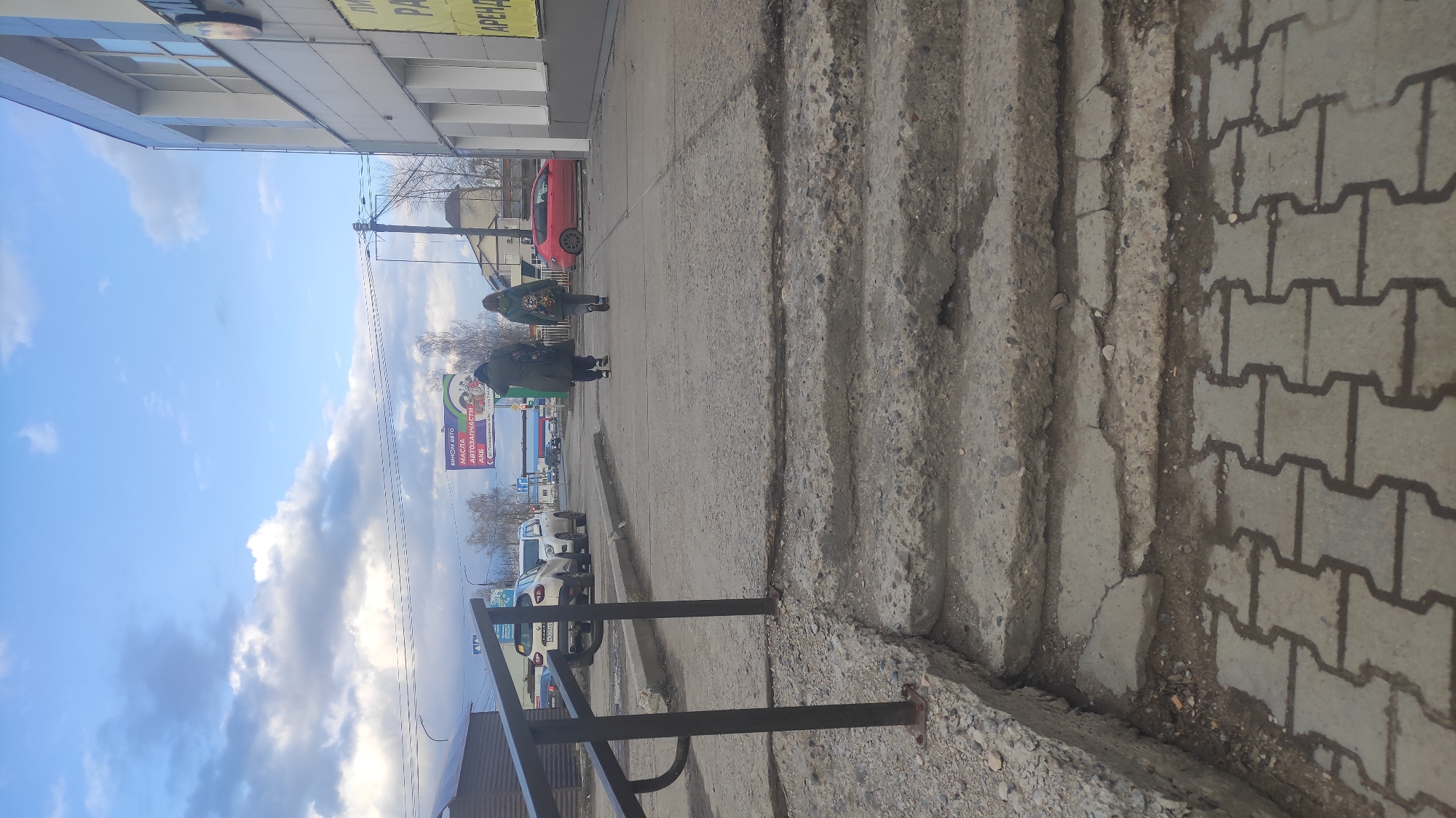 The image shows a street scene in Chaikovsky, Russia. Two people walk along a sidewalk next to a building with a sign in Russian visible in the background. Cars are parked along the street. The sidewalk is made of interlocking concrete pavers, and there are concrete steps leading up to the building. A metal railing is in the foreground. The sky is blue with scattered clouds. A red car is visible further along the sidewalk.