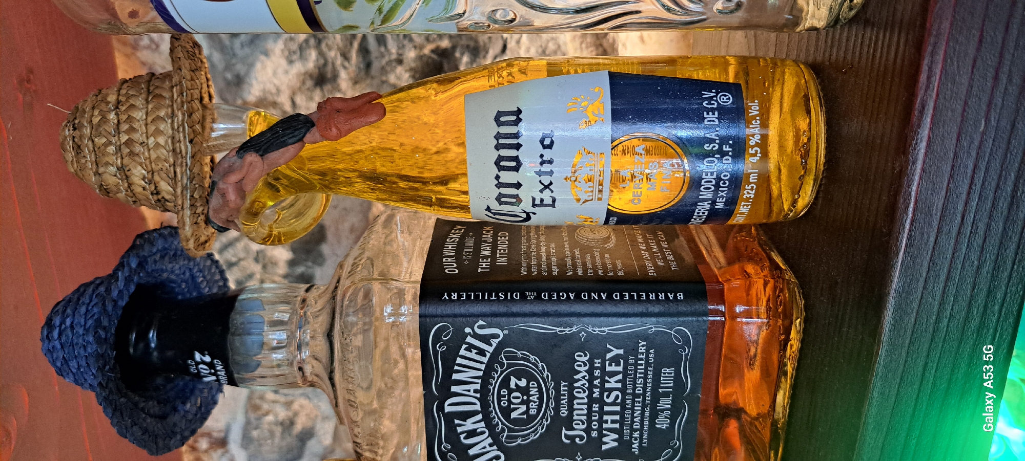 Two bottles of alcohol, a Jack Daniel's whiskey bottle and a Corona Extra beer bottle, are displayed side-by-side on a dark wooden shelf. Both bottles are adorned with small, novelty hats. The Jack Daniel's bottle has a dark blue, woven hat, while the Corona bottle is topped with a light brown, straw hat. A small, figurine hand is visible near the top of the Corona bottle, appearing to hold the straw hat. In the background, a stone wall is visible, suggesting the setting is indoors. The lighting in the foreground appears to include a green hue, possibly from a light source below the shelf. The image is not geo-tagged and therefore cannot be localized to Kos, Greece.