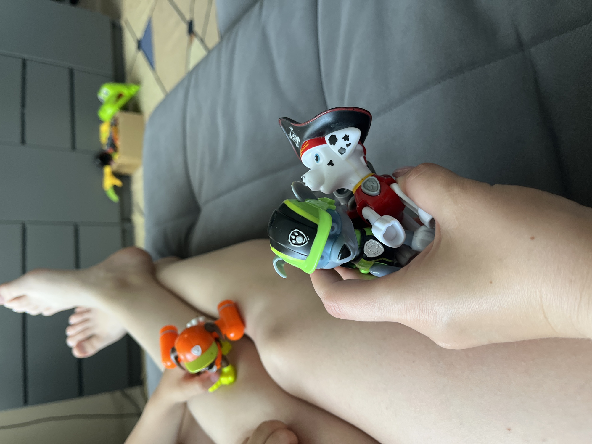 The image depicts a close-up view of a person's legs and hands holding two Paw Patrol toy figurines. The person is seated on a gray couch. In the foreground, two hands are holding a toy figure of a blue dog wearing a green helmet and vest, and a toy figure of a white and black spotted dog dressed as a pirate. Behind the hands, the person's legs are visible, with one leg crossed over the other. A small orange and green toy is also visible on the person's left leg. In the background, a dresser or cabinet and some other toys are partially visible. The scene appears to be indoors, in a domestic setting. The image does not contain any information that allows for its location to be determined as the city of Верхняя Сысерть, Russia.