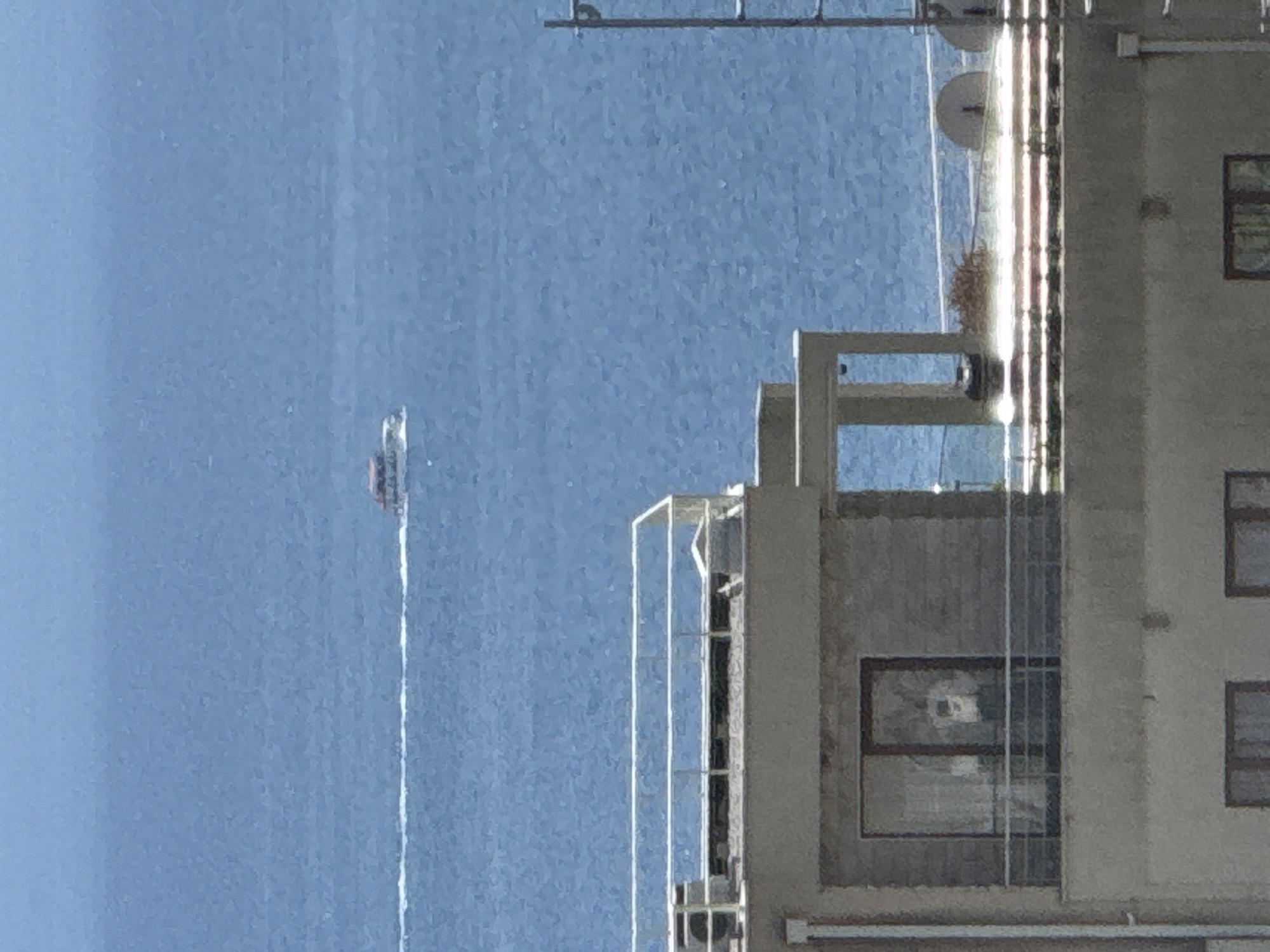 An aerial view captures a wide expanse of blue water, likely the sea, with a distant white wake indicating a boat's movement. In the foreground, a multi-story building dominates the lower portion of the frame. The building's facade is a mixture of light gray and beige concrete. A prominent feature is a rooftop balcony with a clear glass railing. The balcony is furnished with what appears to be satellite dishes and some sparse vegetation. Another building, partially visible at the bottom, has dark-framed windows. The overall scene suggests a coastal urban environment, potentially in Boreti, Montenegro, given the context.