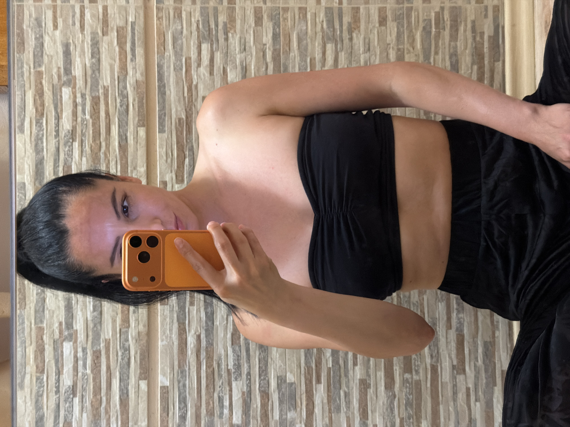 The image depicts a young woman taking a selfie in a bathroom. She has dark hair pulled back in a high ponytail and is wearing a black strapless top and black pants. She is holding an orange smartphone in her right hand, which is angled to capture her reflection in the mirror. The background is a tiled wall with horizontal rows of rectangular tiles in various shades of brown and gray. The lighting appears to be artificial and casts a slight sheen on her skin. The location is identified as a bathroom in Pleven, Bulgaria.