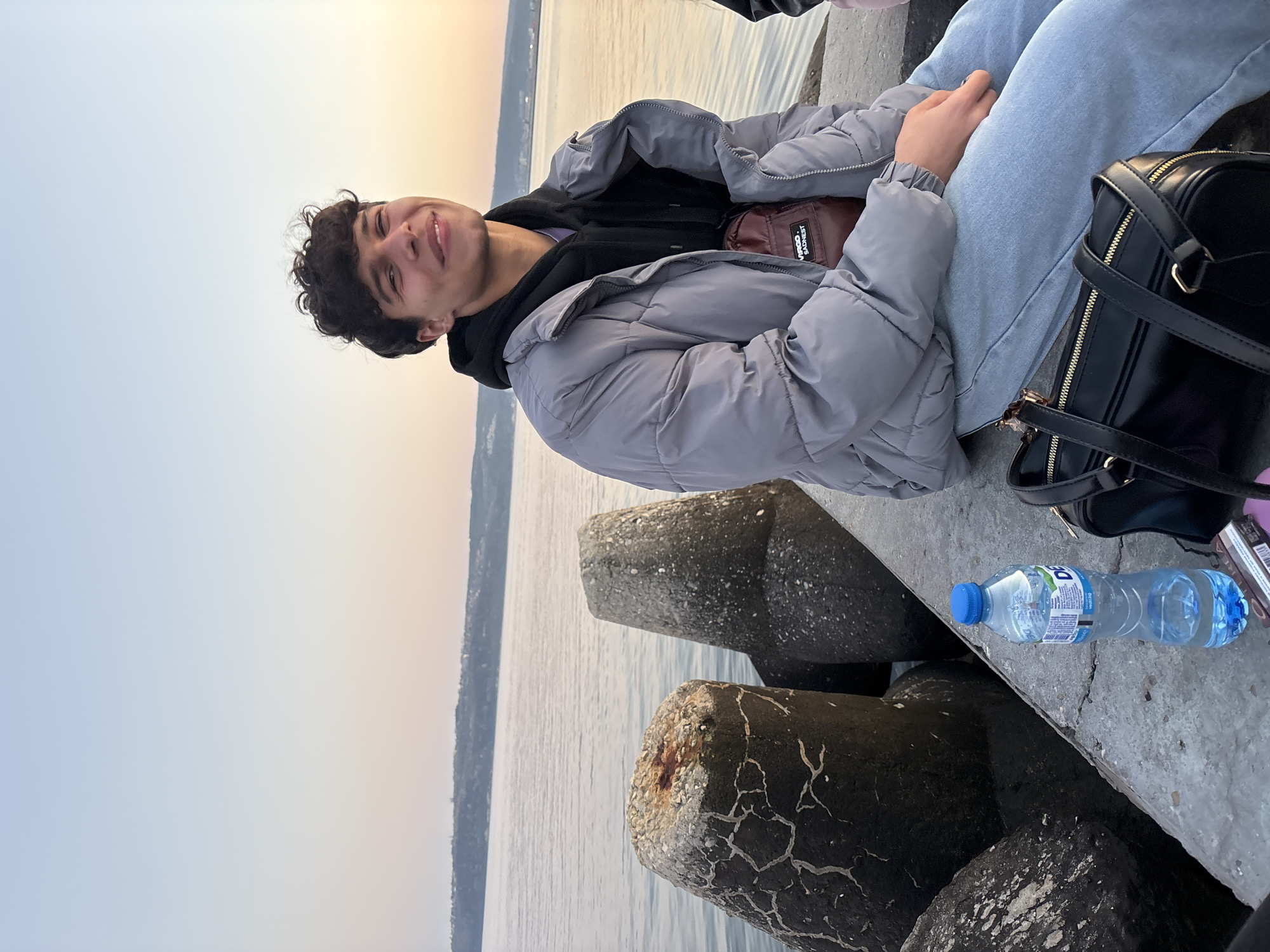 A young person is sitting on a concrete pier near a body of water. The person has dark curly hair and is smiling. They are wearing a gray puffer jacket over a black hoodie and light blue jeans. A black backpack is next to them. A plastic water bottle is placed on the concrete surface. In the background, the water meets the sky at the horizon. The city is Unknown, Unknown.
