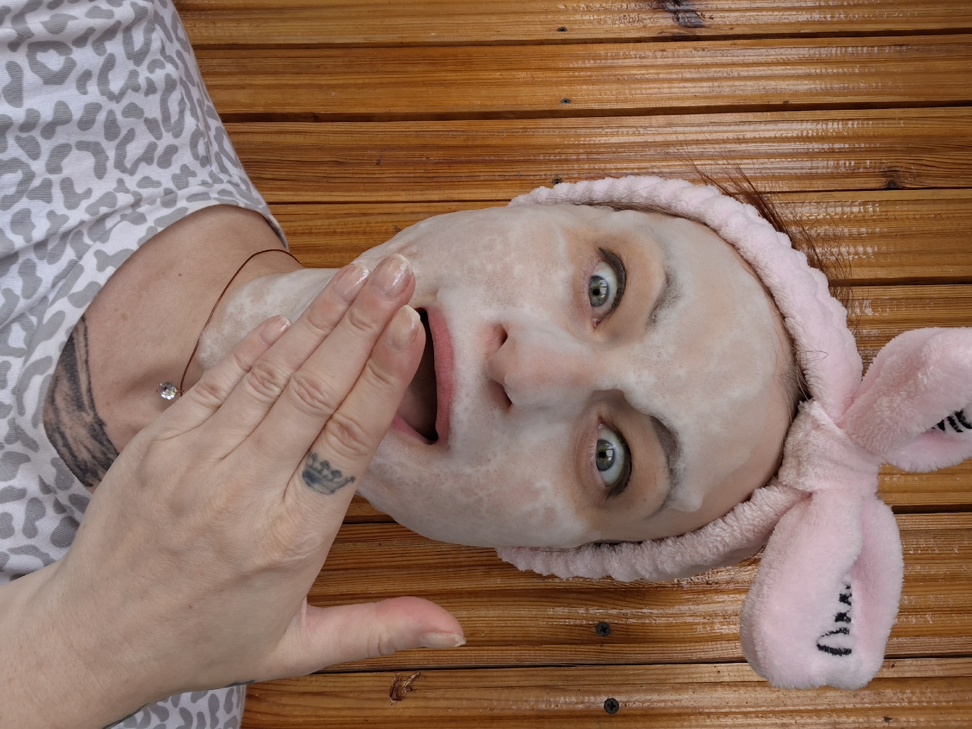 A medium shot captures a person with a white facial mask on, wearing a pink fuzzy headband with rabbit ears, and a gray and white leopard print shirt. The person's eyes are wide with surprise or excitement, and their mouth is open, with a hand covering the lower half of their face. The background consists of vertical wooden planks, likely part of an outdoor structure. There is a small diamond pendant visible on a thin necklace. Tattoos are visible on the person's hand and neck. The lighting is natural, suggesting an outdoor setting. The overall scene appears to be a candid moment, possibly captured during a skincare routine or a playful pose. There is no information about the specific location within Забалоцкі сельскі Савет, Belarus.