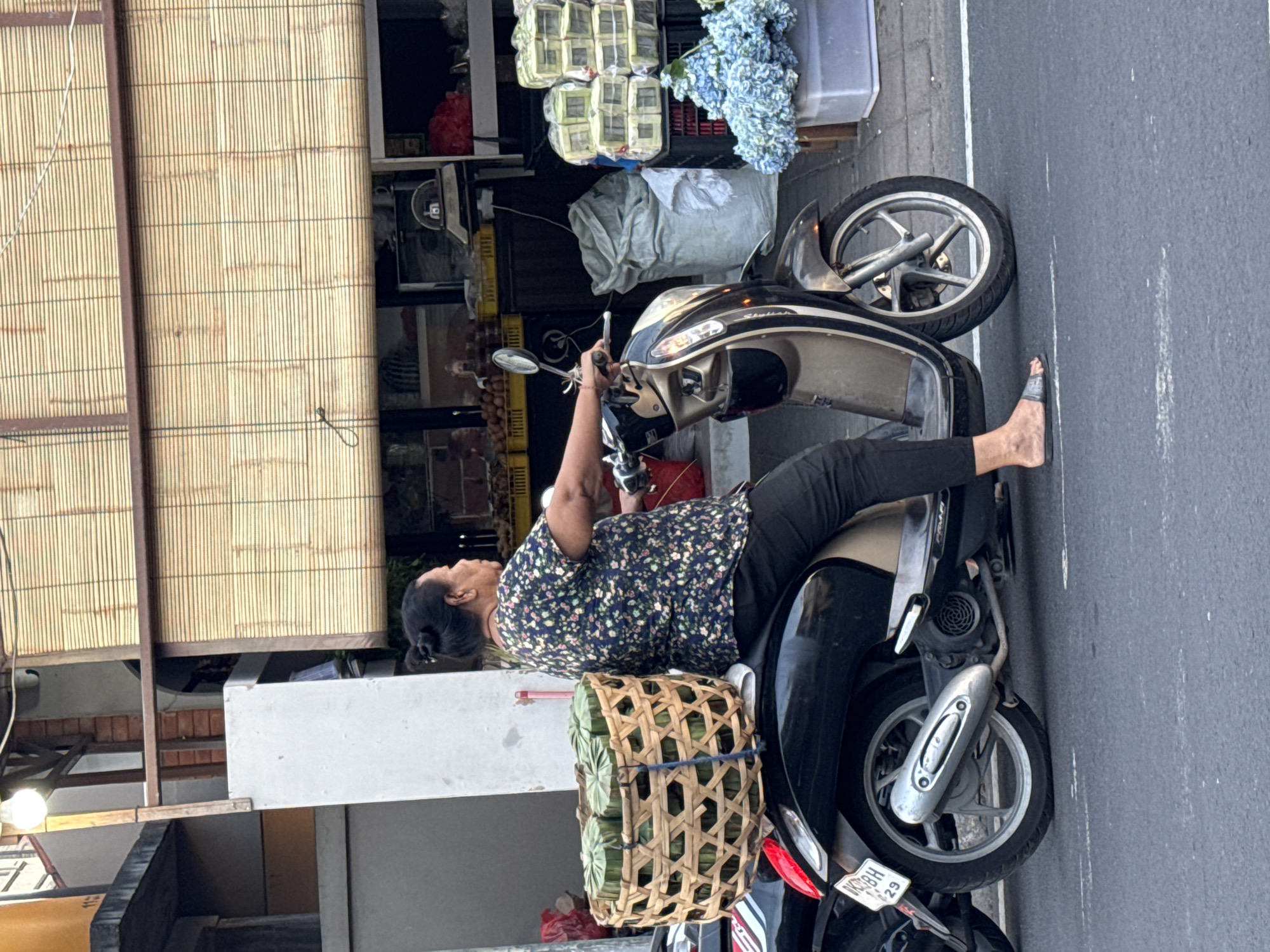 An Indonesian woman with her hair in a bun is riding a scooter in Kuta, Indonesia. She is wearing a floral print shirt and black pants. The scooter is carrying a large woven basket filled with green cylindrical items, possibly food wrapped in leaves, secured to the back. In the background, there is a storefront with a bamboo blind, shelves stocked with goods, and blue flowers in a large container. The scooter is on a paved road with a white line marking the lane.