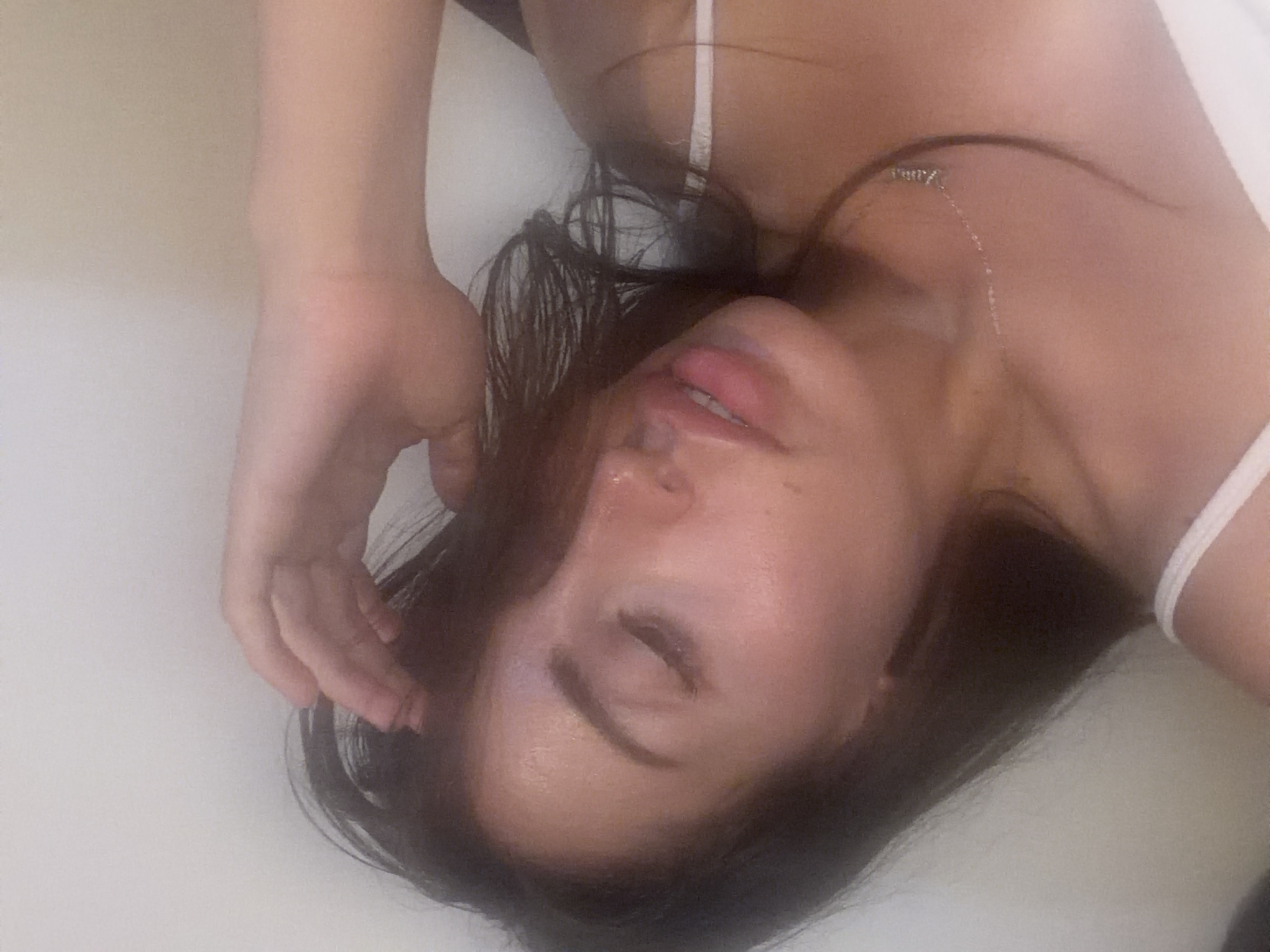 A close-up, high-angle shot captures a young woman lying down, her head tilted back and a hand resting on her dark hair. Her eyes are closed, and her lips are slightly parted. A thin necklace with a nameplate is visible around her neck. She is wearing a white strap top. The background is a plain, pale wall. The lighting is soft and diffused, with a warm glow. The scene is intimate and relaxed. The image is not geo-tagged, so the specific location in Tersefanou, Cyprus, cannot be determined.