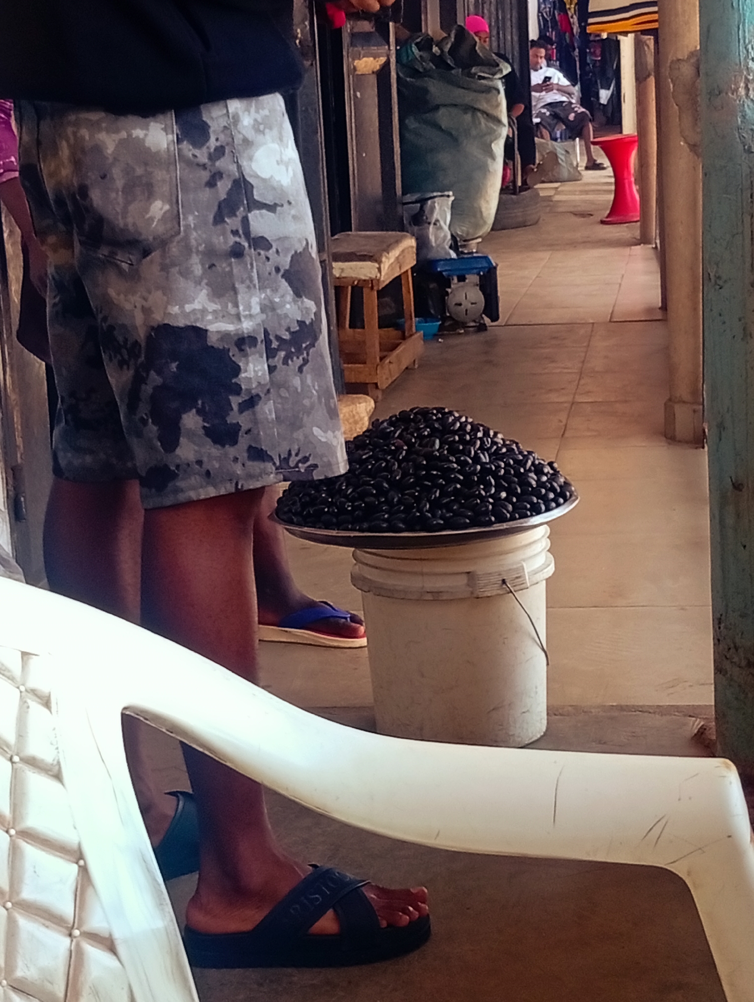The image captures a street scene in Bukuru, Nigeria. In the foreground, a person wearing camouflage shorts and black sandals is standing. In front of them is a white plastic chair. To the right of the person, a white bucket supports a metal tray filled with black beans. In the background, other individuals are visible. One person in a pink hat is standing near a large green bag. Another person is seated in a white shirt and shorts, looking at their phone. A bright red stool is also present in the background. The environment appears to be an outdoor market or walkway with tiled flooring.