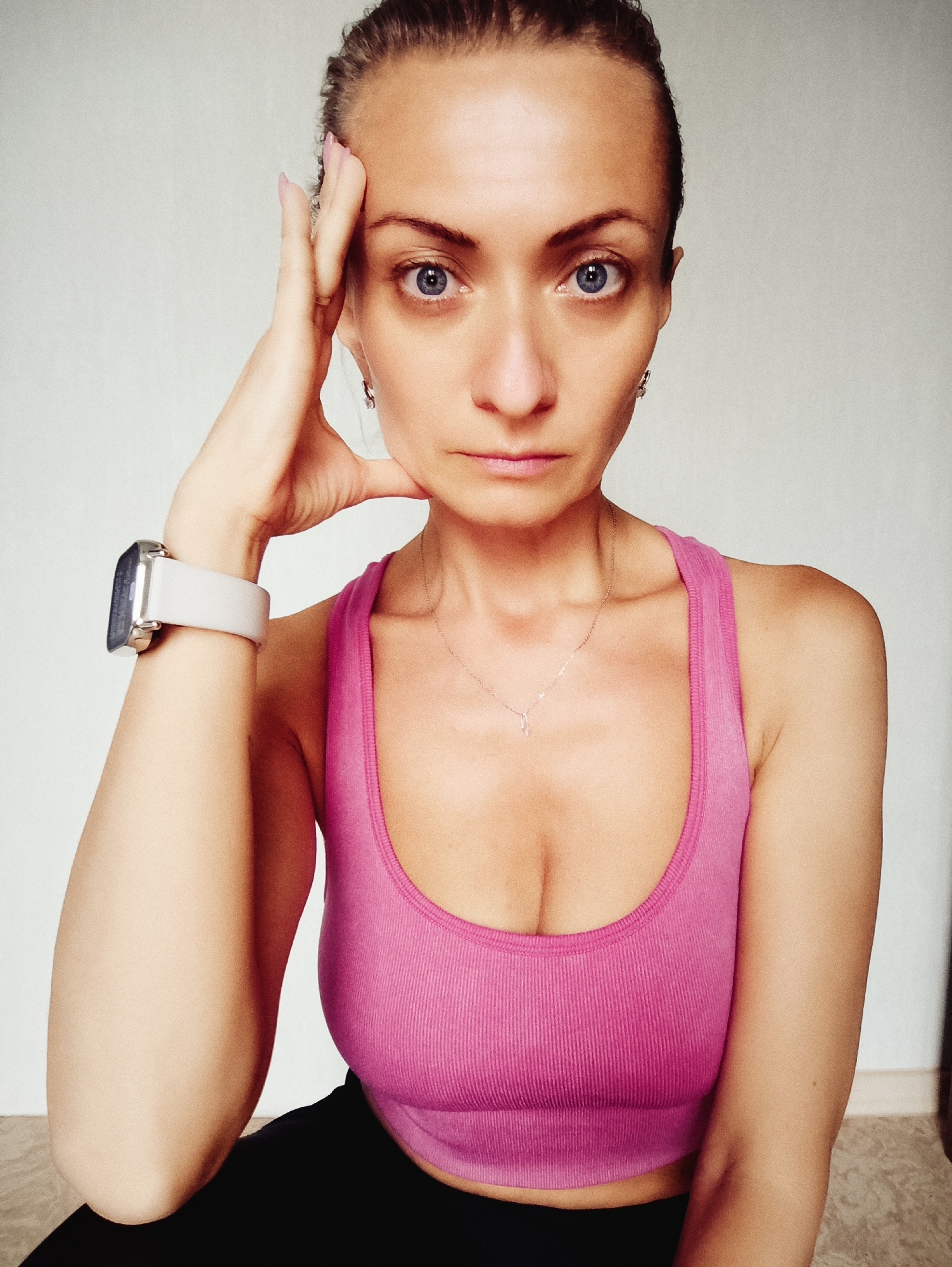 A woman is posing for a selfie in what appears to be a domestic setting, possibly an apartment in Moscow, Russia. She has her left hand on her forehead and is looking directly at the camera with a serious expression. She is wearing a pink ribbed sports bra and black bottoms. A light-colored smartwatch is visible on her left wrist. The background is a plain, light-colored wall with a slight texture and a sliver of a floor visible at the bottom. The lighting suggests an indoor environment, possibly near a window.