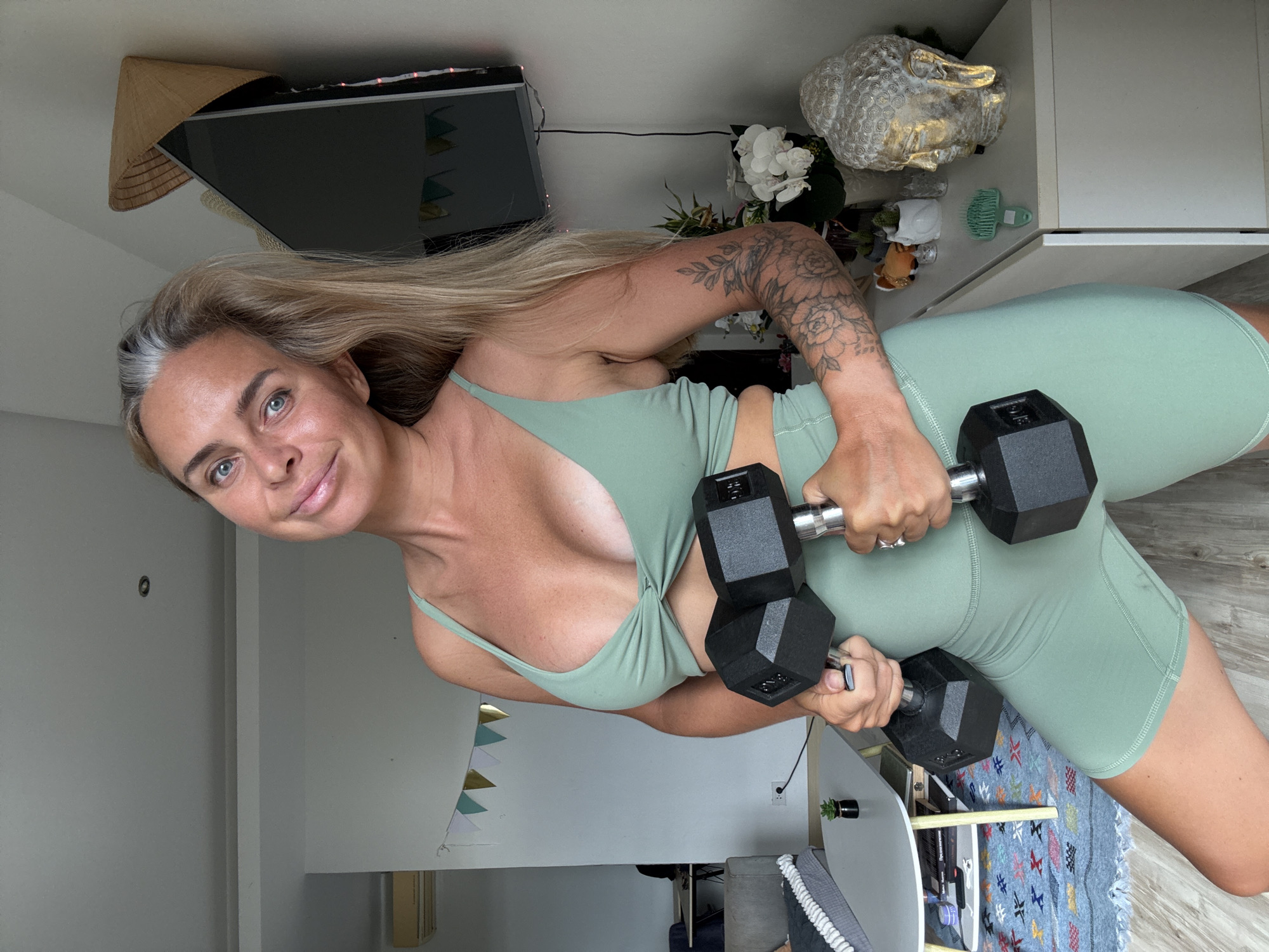 A woman with blonde hair and blue eyes stands in an indoor setting, holding dumbbells. She is wearing a mint green sports bra and shorts. Her left arm is tattooed with floral designs. In the background, there is a television, a straw hat, a Buddha statue, and various decorative items. The room appears to be a living space, with furniture and a rug visible. The lighting suggests an indoor environment.
